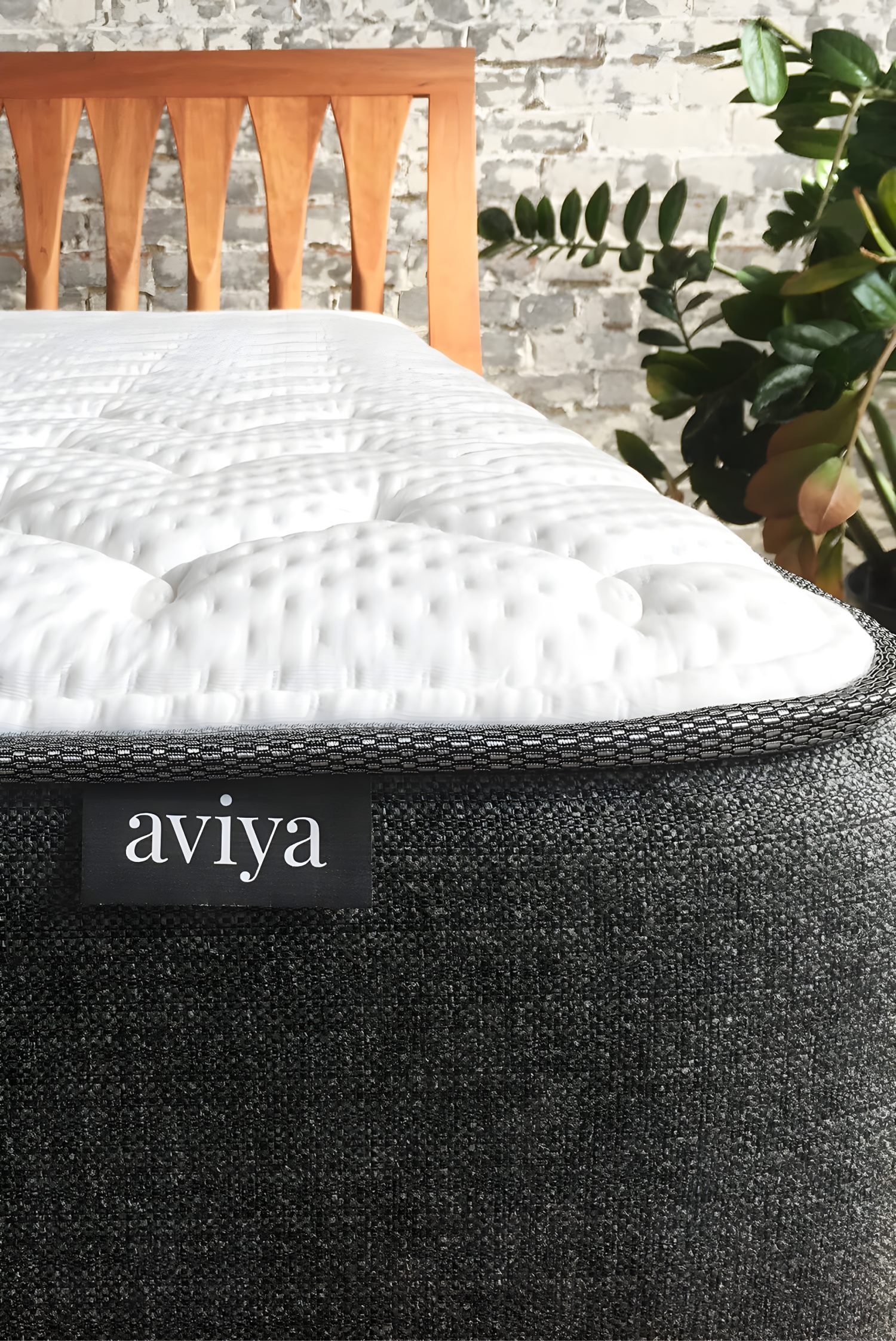 Aviya mattress close-up detail on wood bed frame, showing textured gray fabric and white quilted top against brick wall