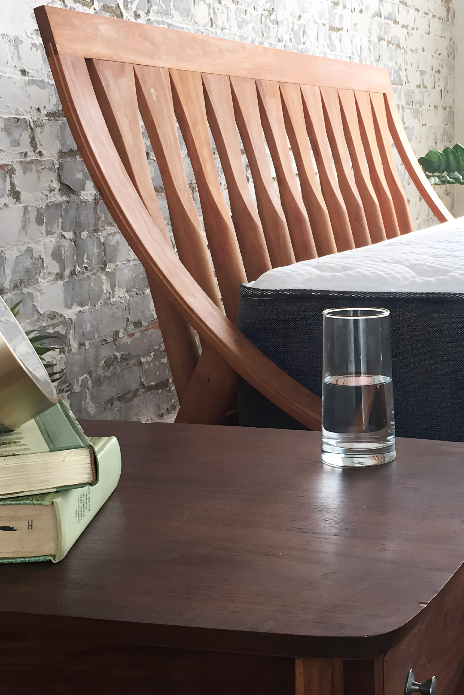 Close-up of wooden bed frame and nightstand with glass of water, books, and exposed brick wall styled for photography