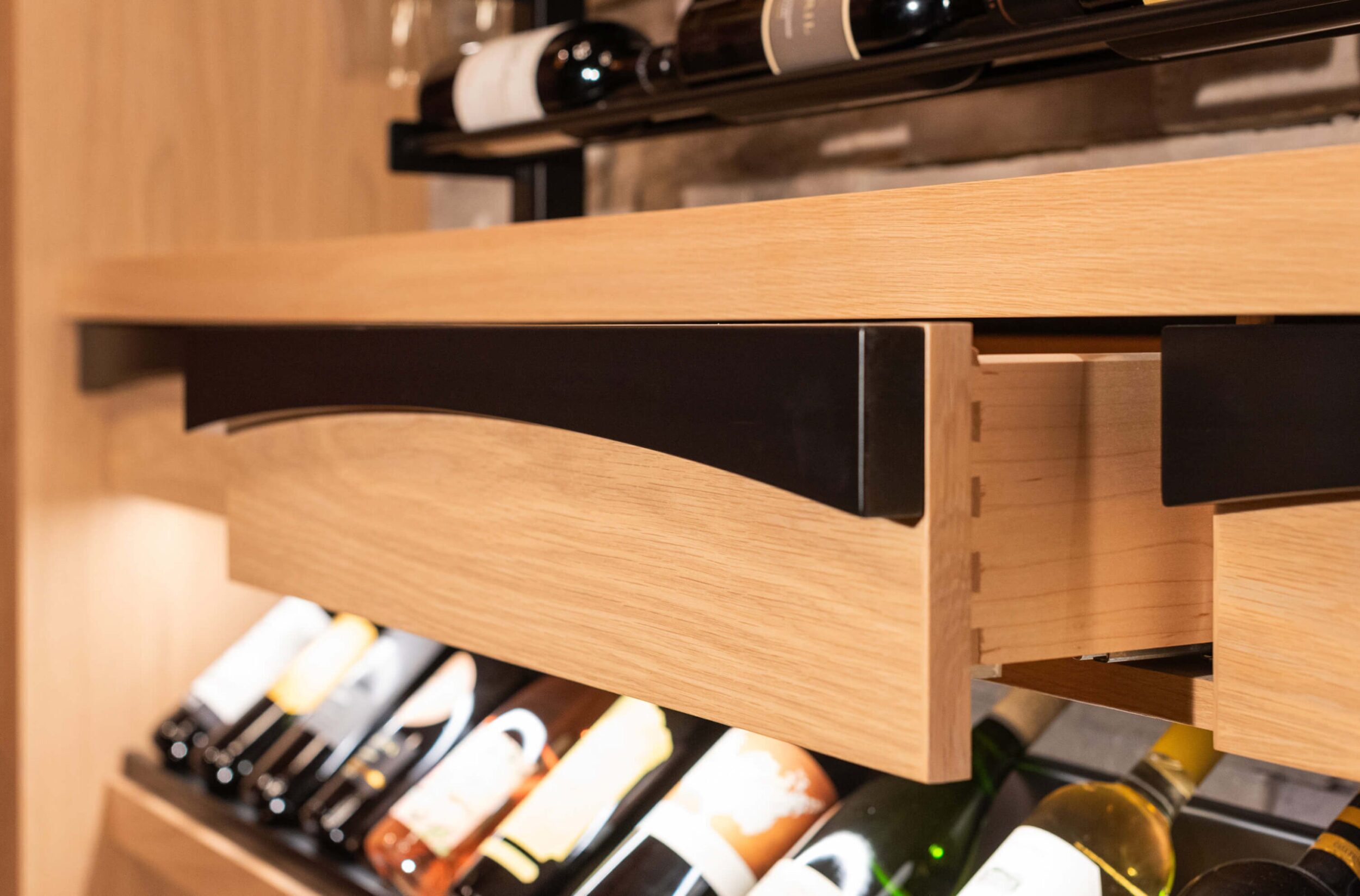 Open wooden drawer under wine rack, showcasing dovetail joints, with illuminated bottles of wine stored below shelf