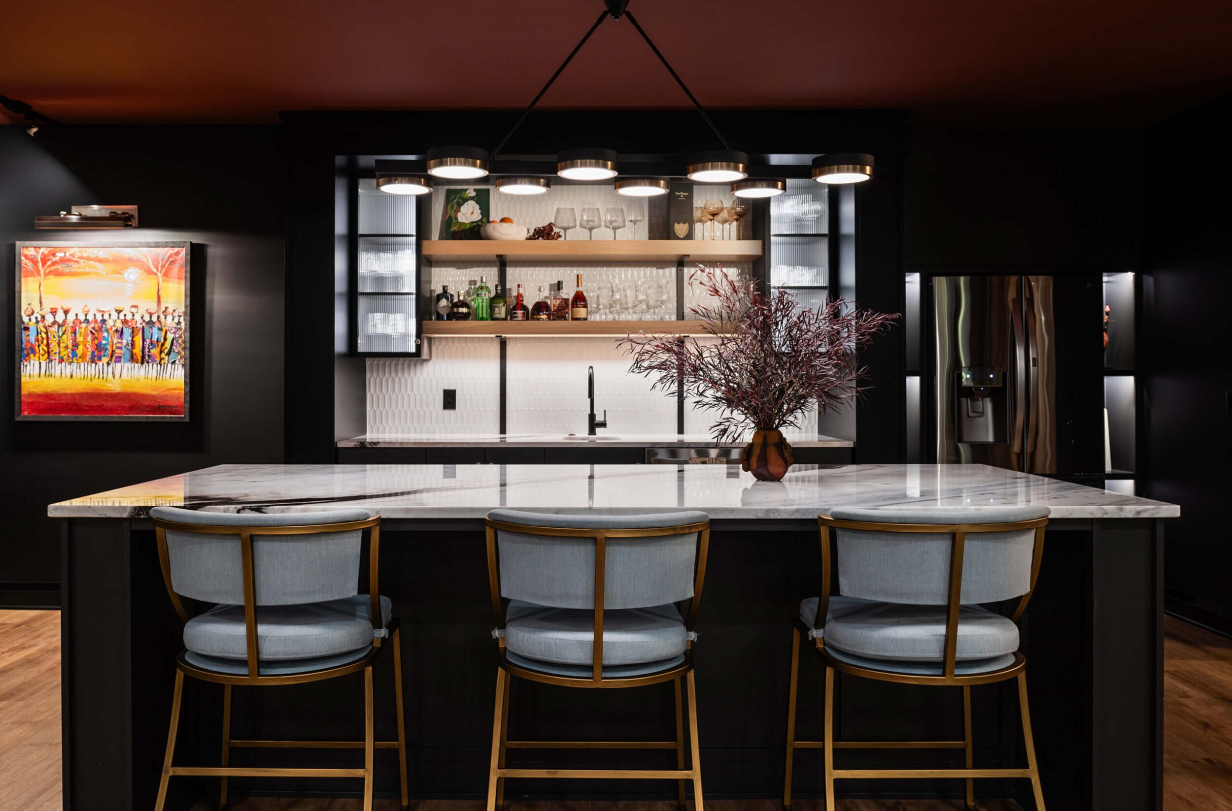 Stylish home bar with marble counter, three cushioned stools, wall shelves of liquor bottles, and bold artwork accent piece