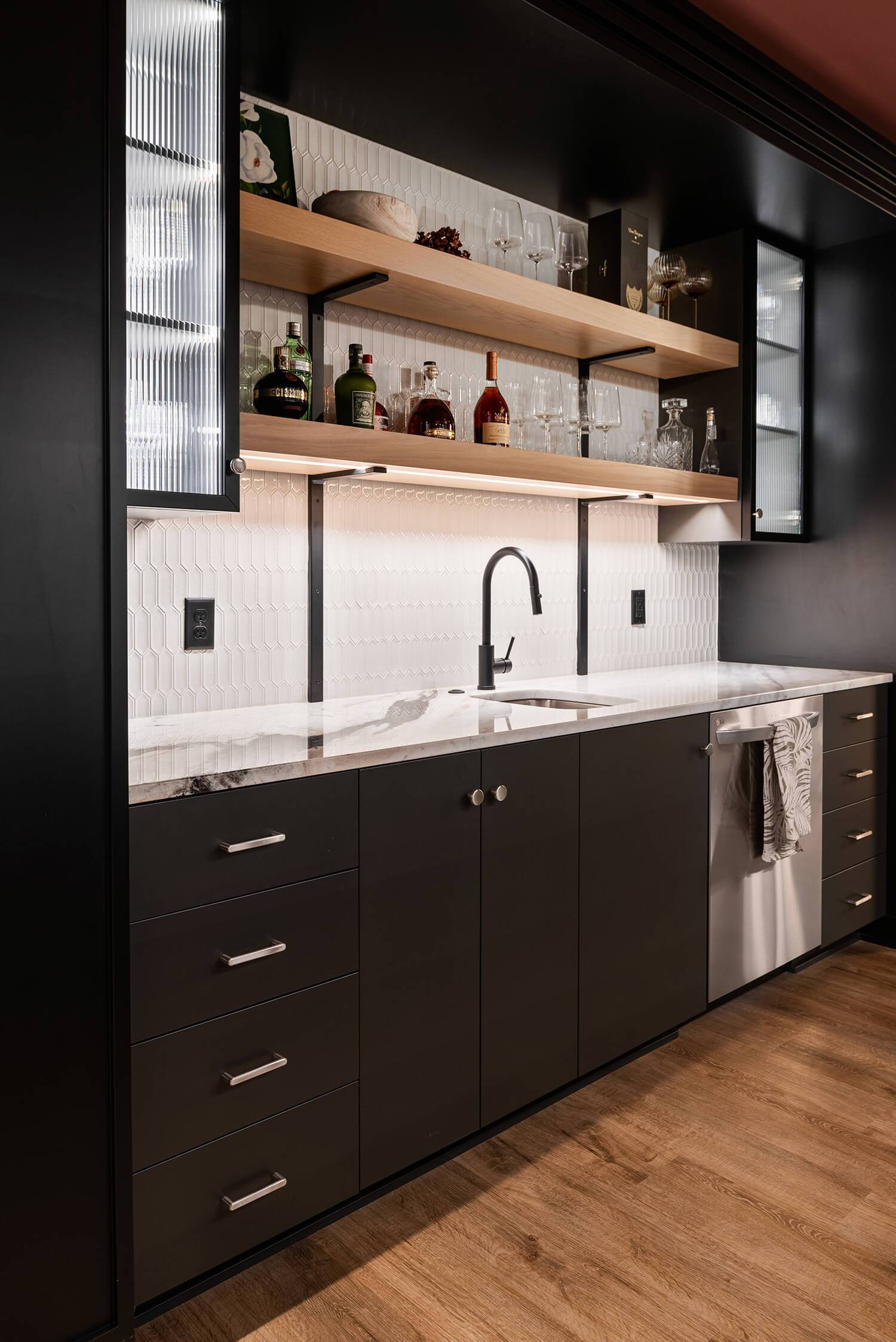 Modern wet bar with black cabinetry, marble counter, open shelves with bottles and glasses, sleek faucet and white backsplash