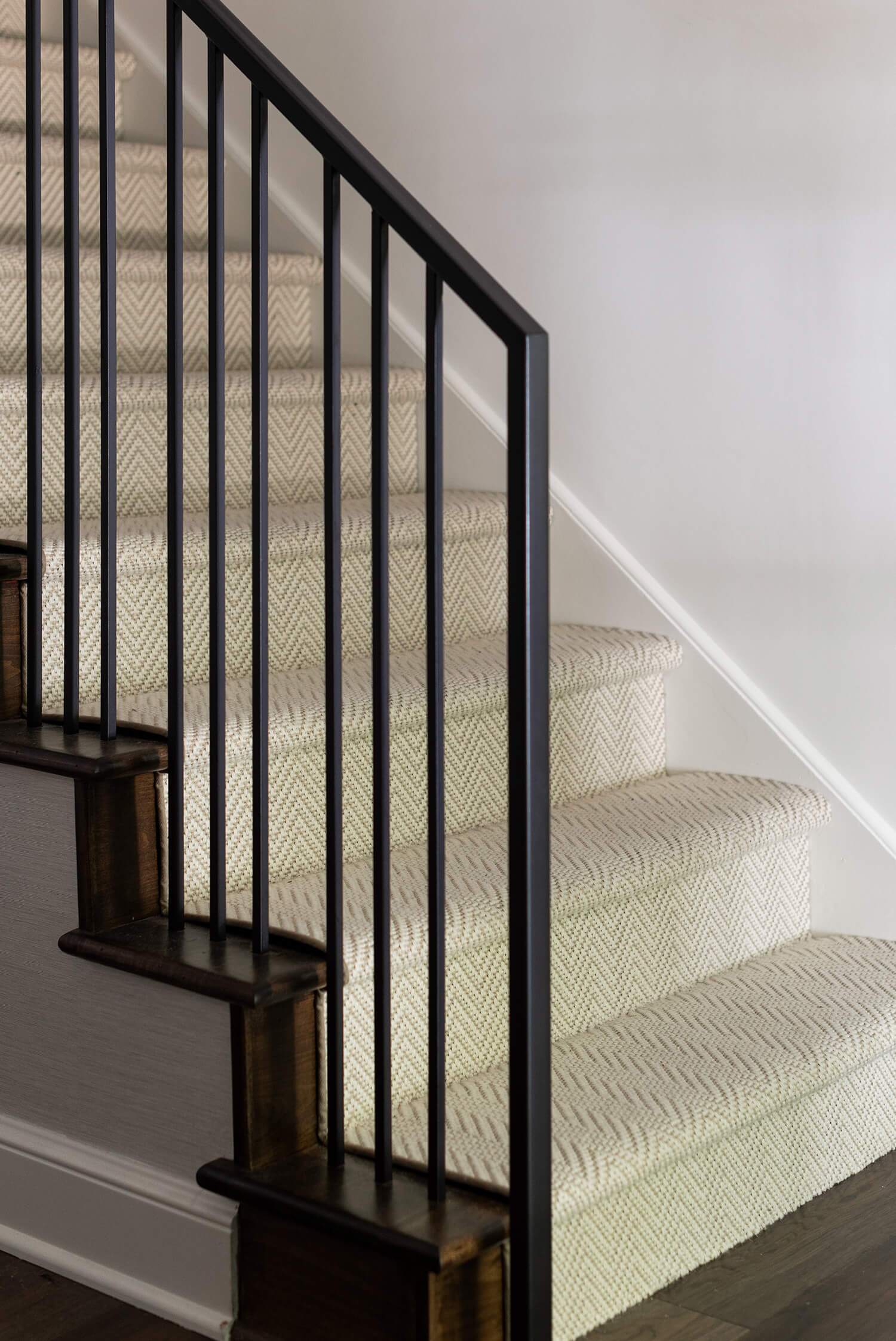 Staircase with cream patterned carpet runner and black metal railing against white wall.