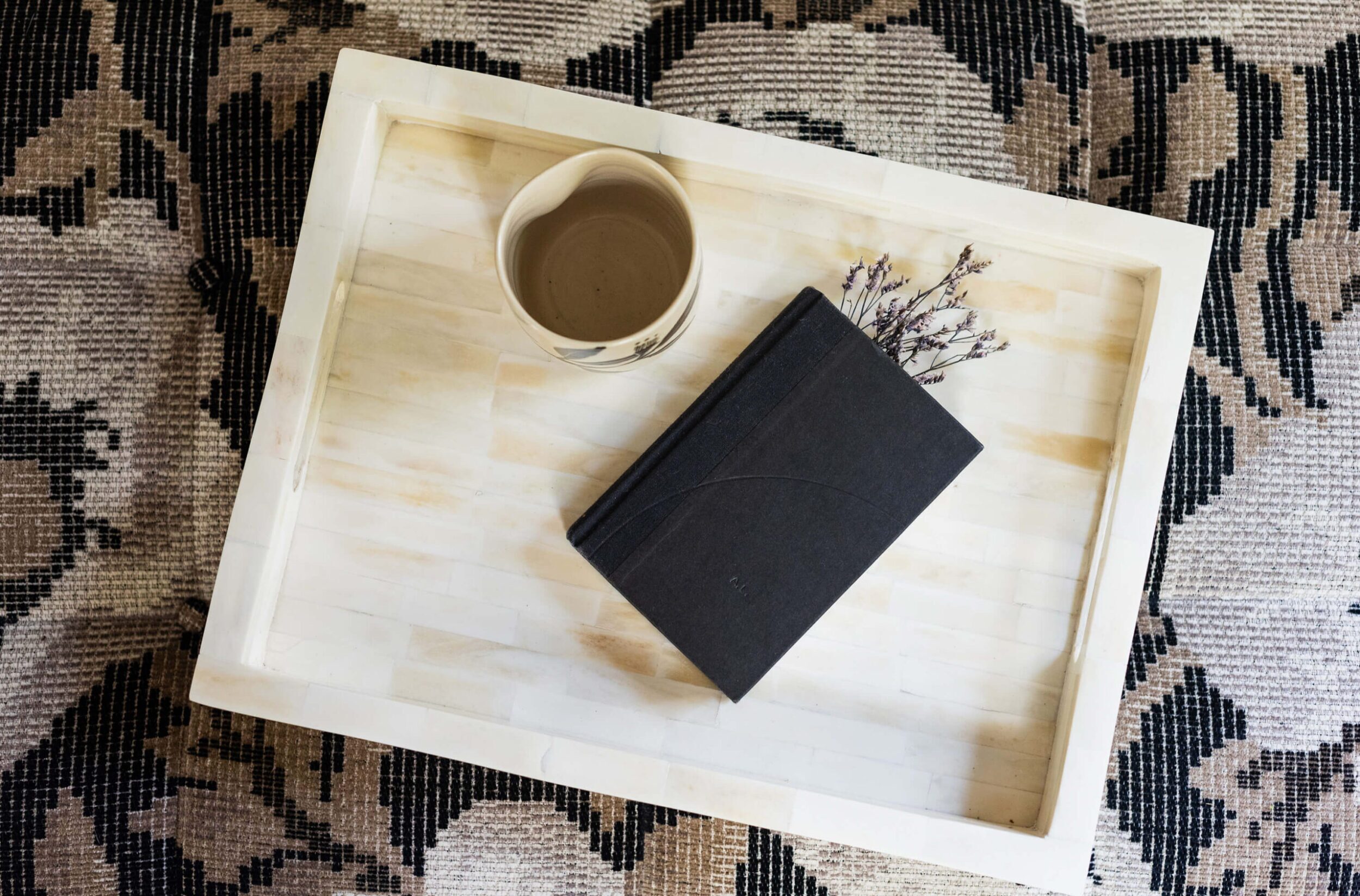 White tray on patterned rug holding a dark book, small ceramic cup, and sprig of lavender.