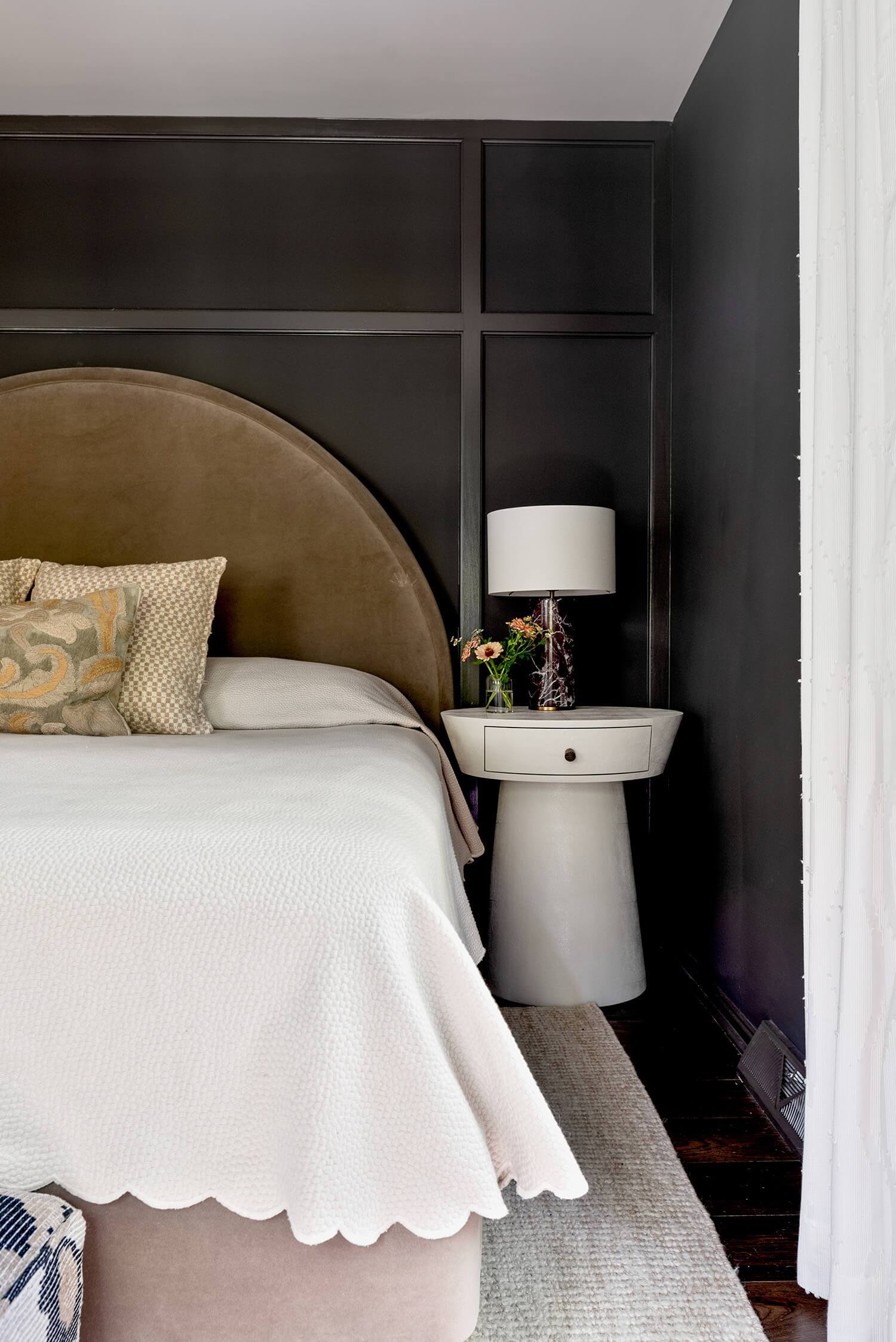 Bedroom corner with round white nightstand, lamp, and fresh flowers beside upholstered bed.
