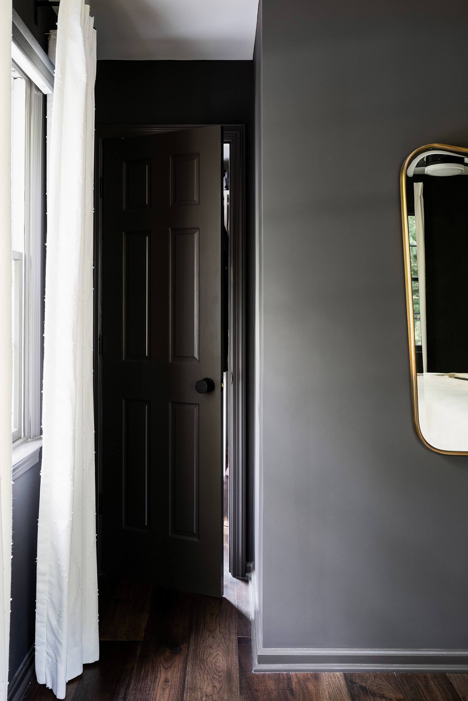 Dark hallway with black door, gold-edged mirror, and white curtain framing a window.
