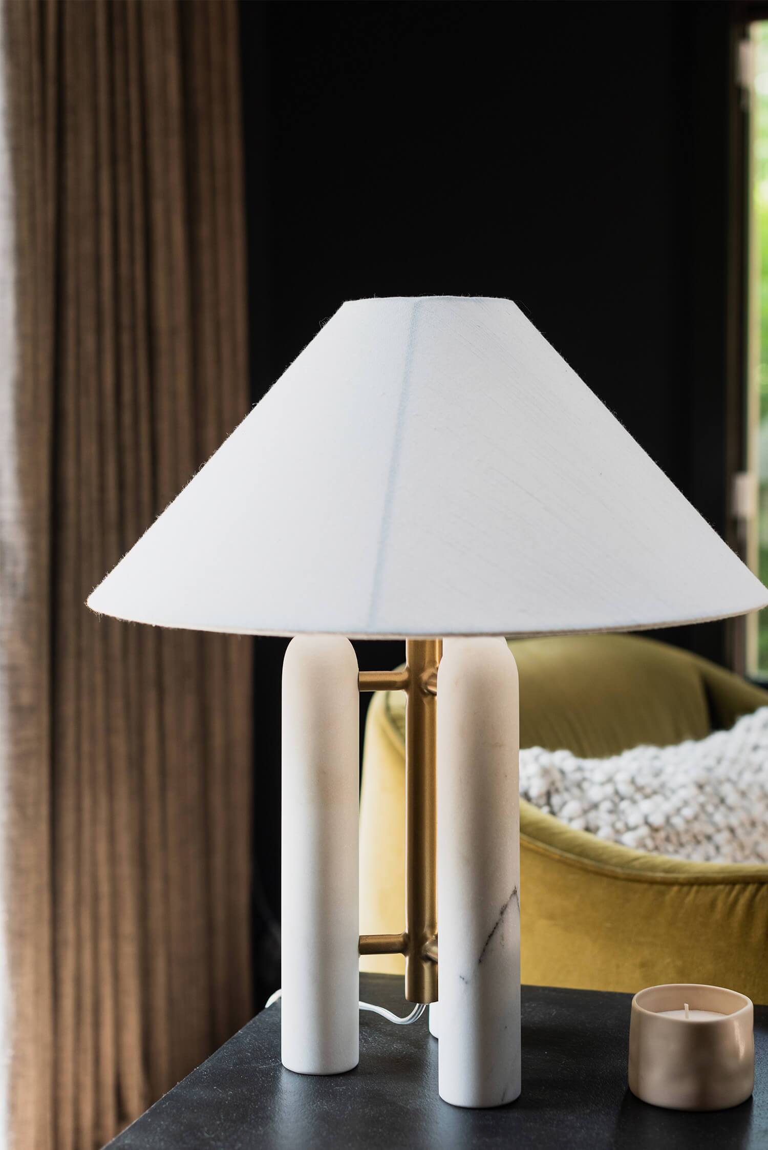 Close-up of modern white table lamp with conical shade on dark side table beside olive green armchair.