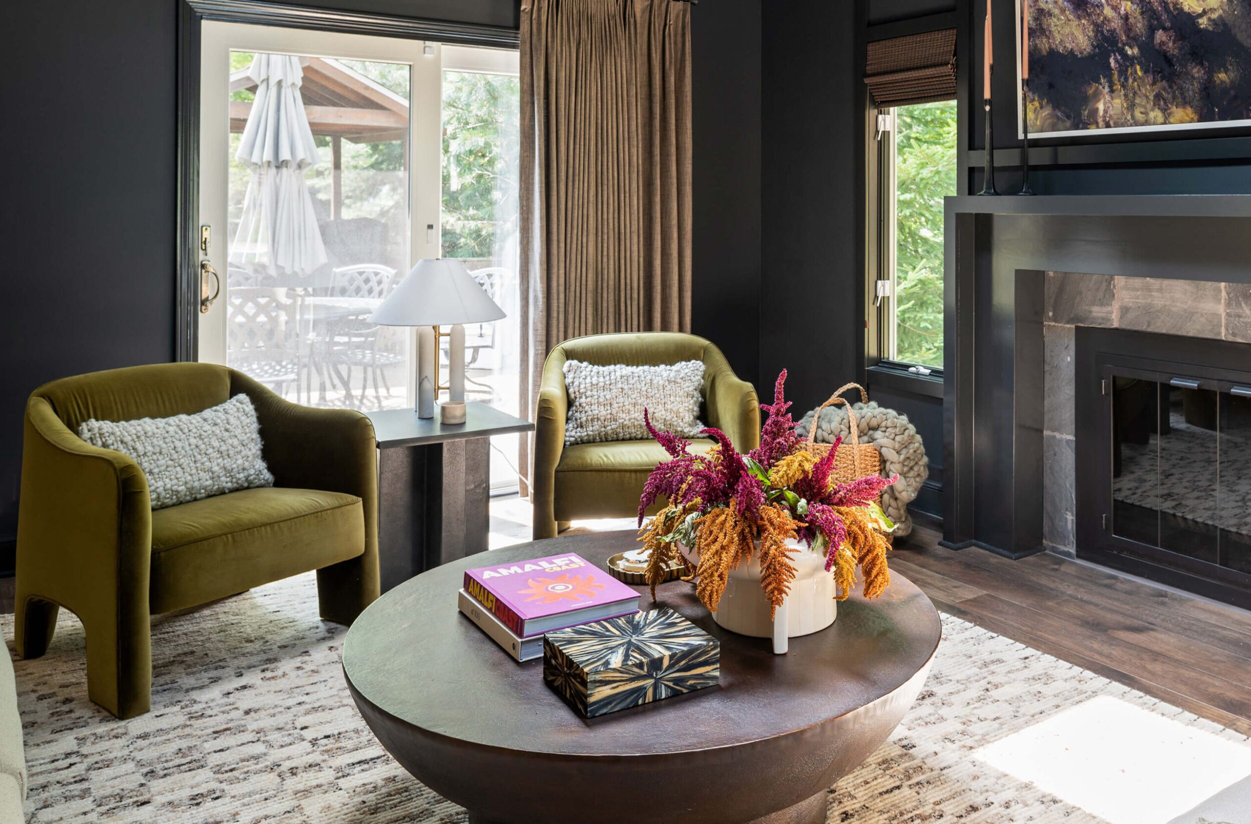 Living room with green velvet chairs, round coffee table, fresh flowers, and dark walls with large windows.