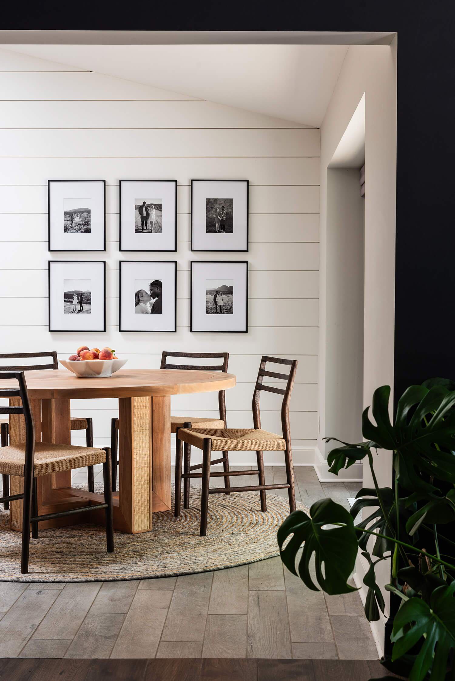 Dining nook with round wooden table, woven chairs, and black-and-white photo gallery wall on white shiplap.