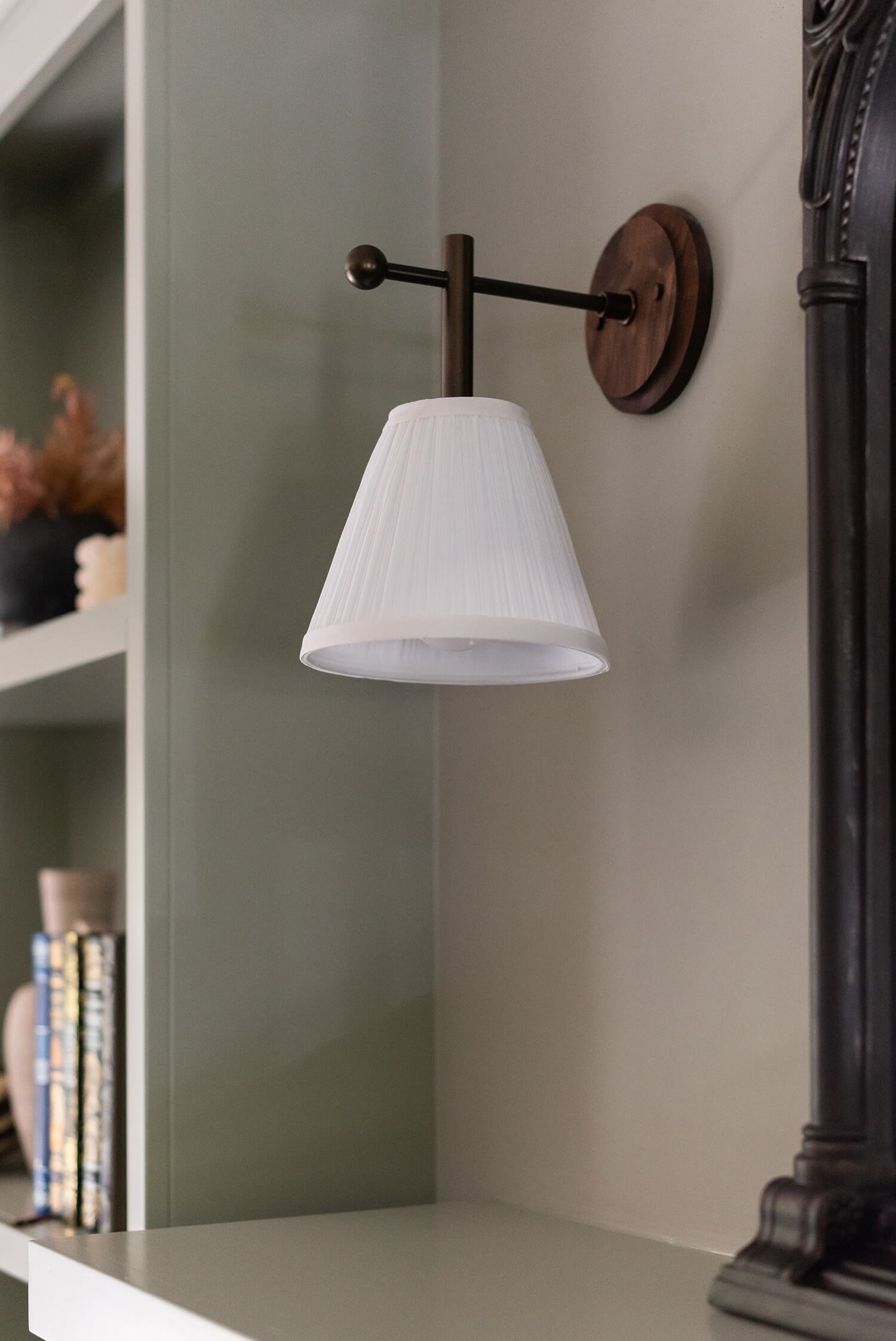 Elegant wall-mounted sconce with white pleated shade and wood backplate, styled in a sage built-in living room corner.