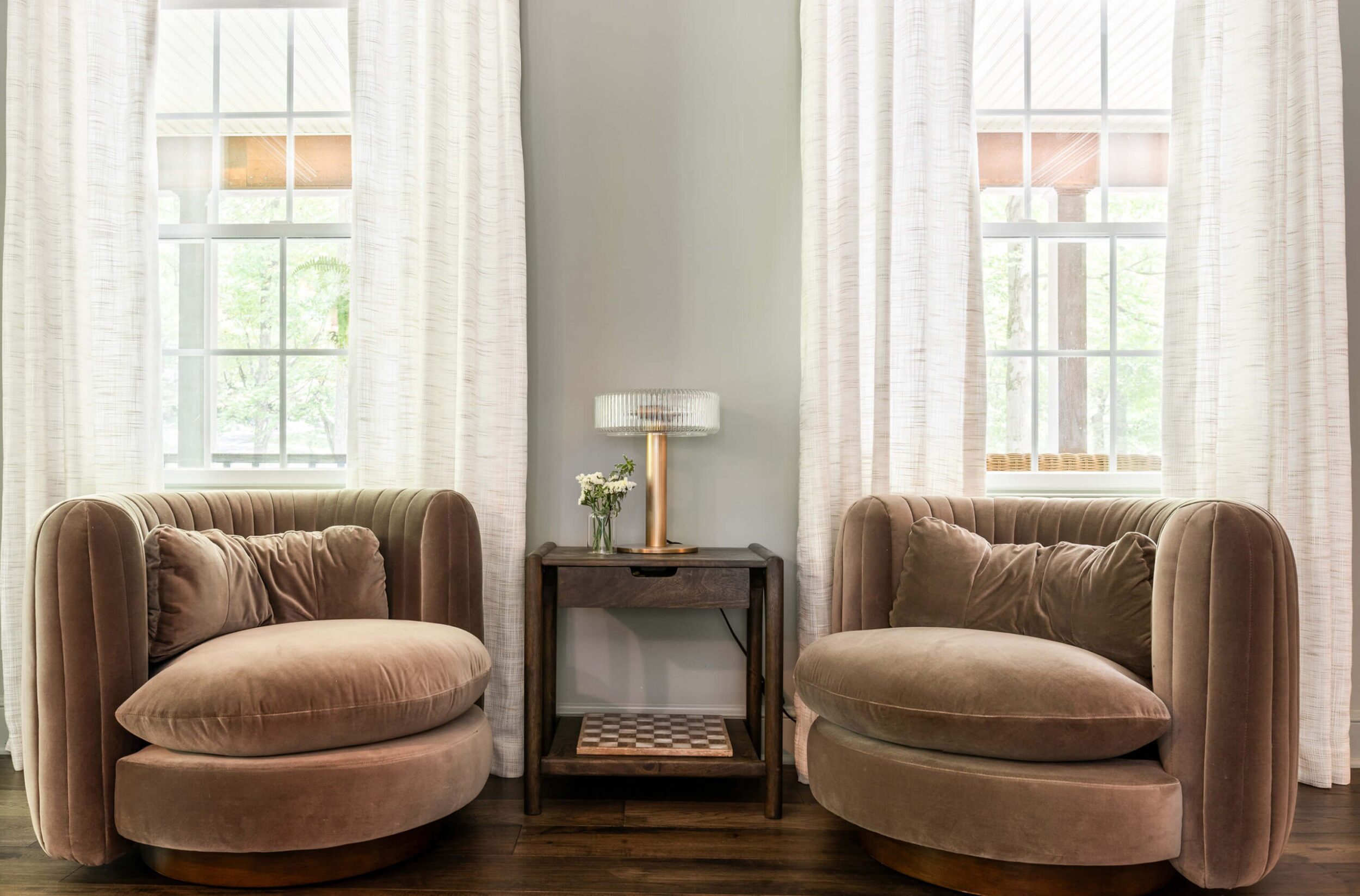 Cozy seating area with plush taupe swivel chairs, side table with lamp and flowers, framed by tall windows with white drapes.
