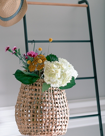 Woven basket with fresh hydrangea, yellow billy balls, and pink flowers hanging on a black ladder with hat above