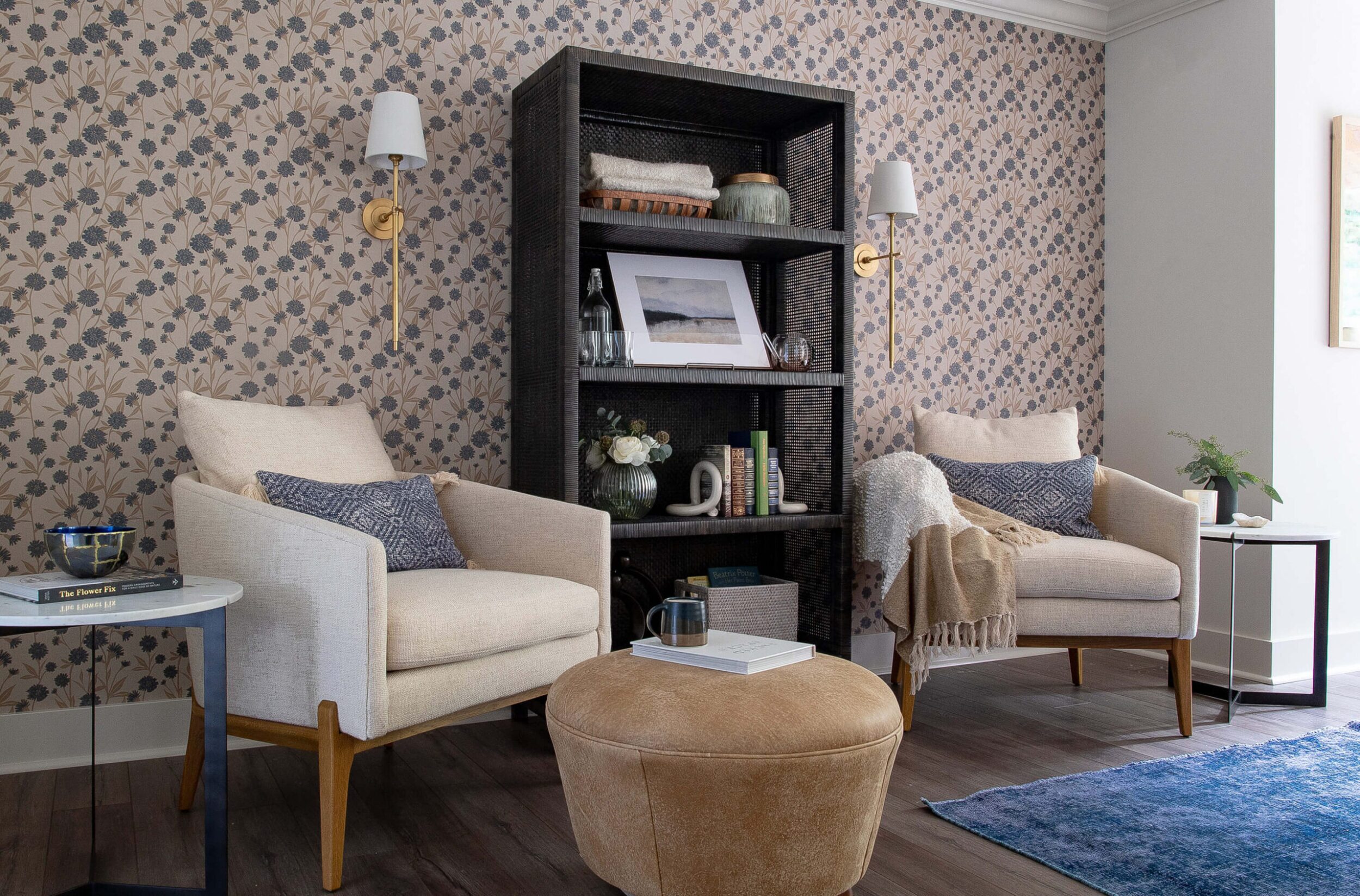 Living space with floral wallpaper, two cream armchairs, ottoman, bookshelf, and warm gold sconces for a cozy atmosphere.