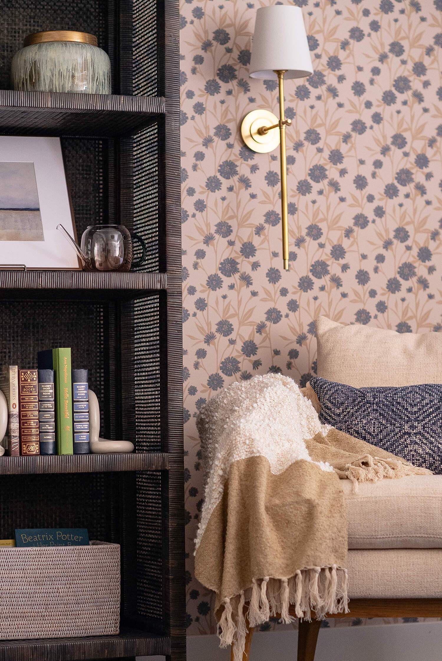 Cozy reading nook with patterned wallpaper, armchair, throw blanket, bookshelf with decor, and brass wall sconce lighting.