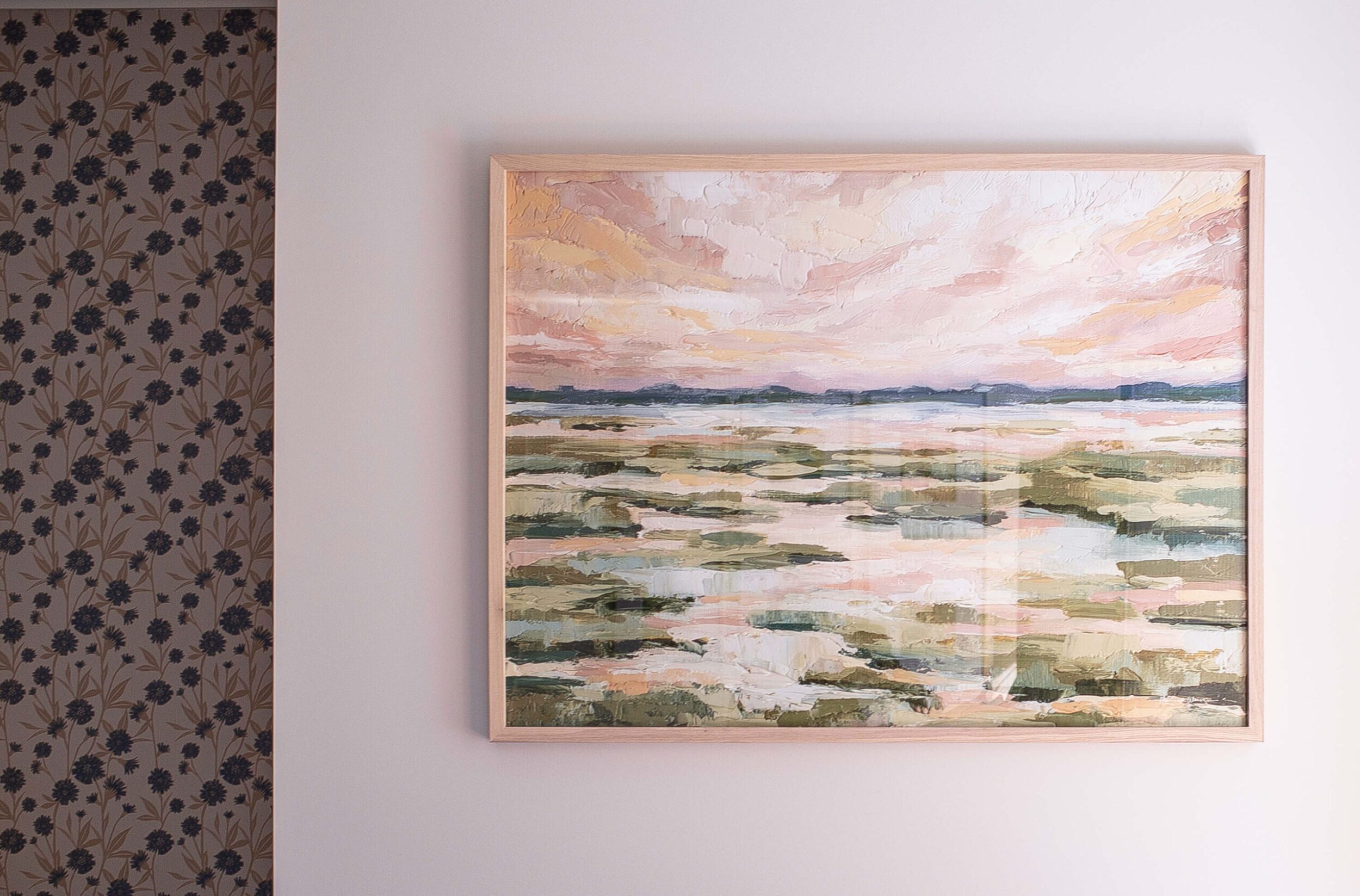 Framed abstract landscape artwork with warm pastel tones hangs beside floral wallpaper, adding charm to the bedroom wall.