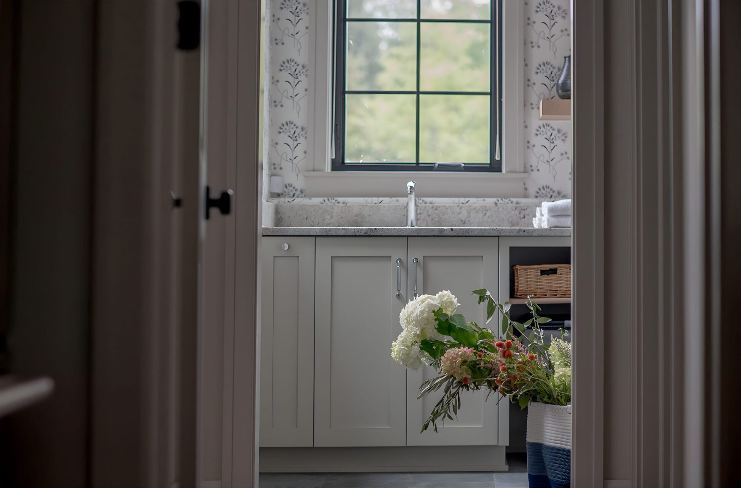 Stylish laundry room sink area with gray cabinetry, window above, woven basket, and floral arrangement in bright setting.
