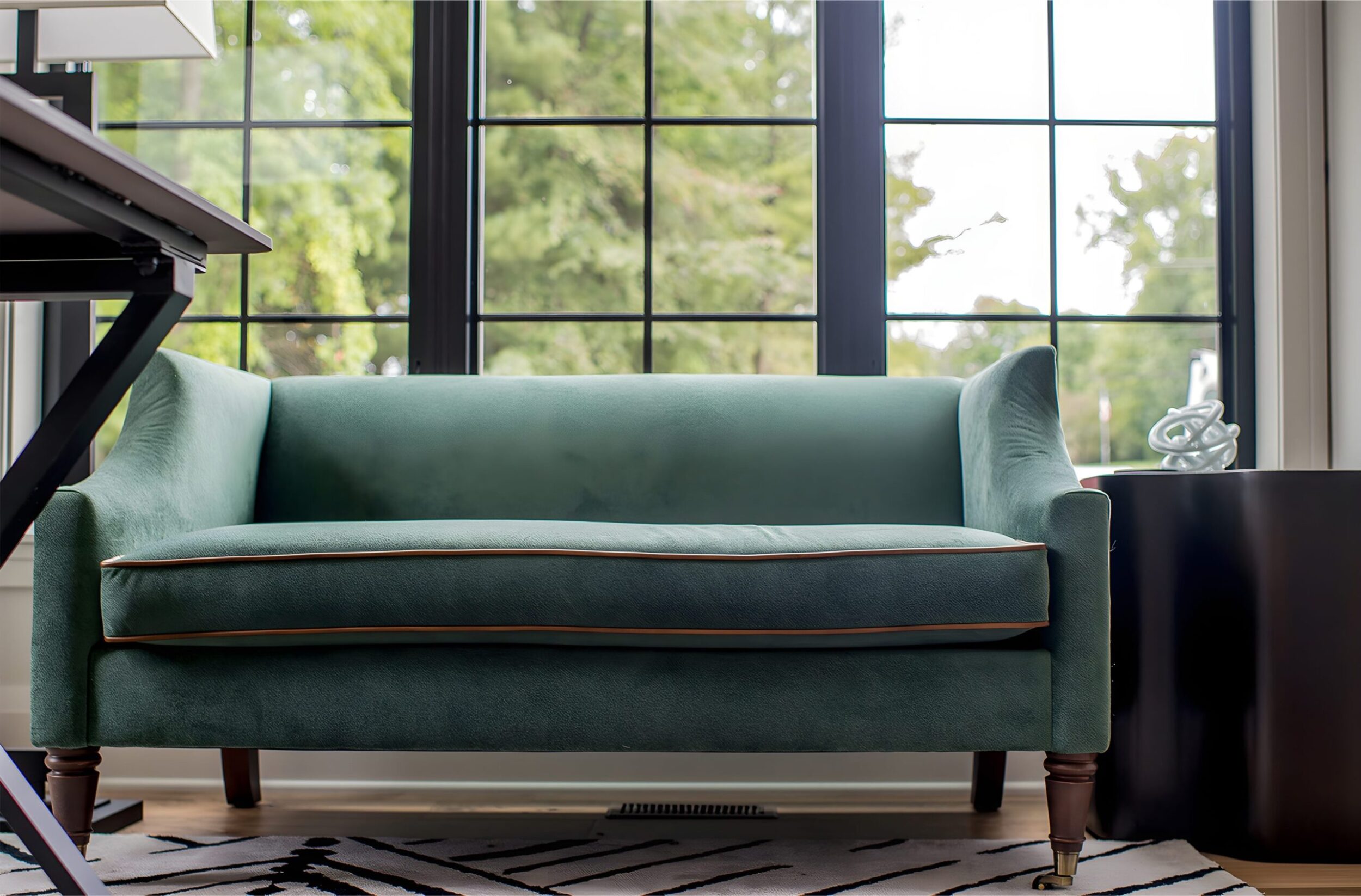 Green upholstered sofa with wood legs placed in front of large grid windows, beside black side table with sculpture