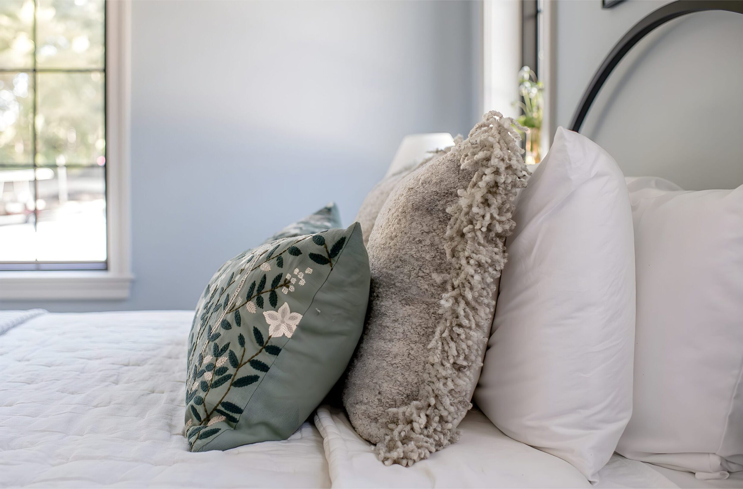 Side view of neatly styled bed with layered pillows, textured gray cushion, and soft white bedding in a serene room.