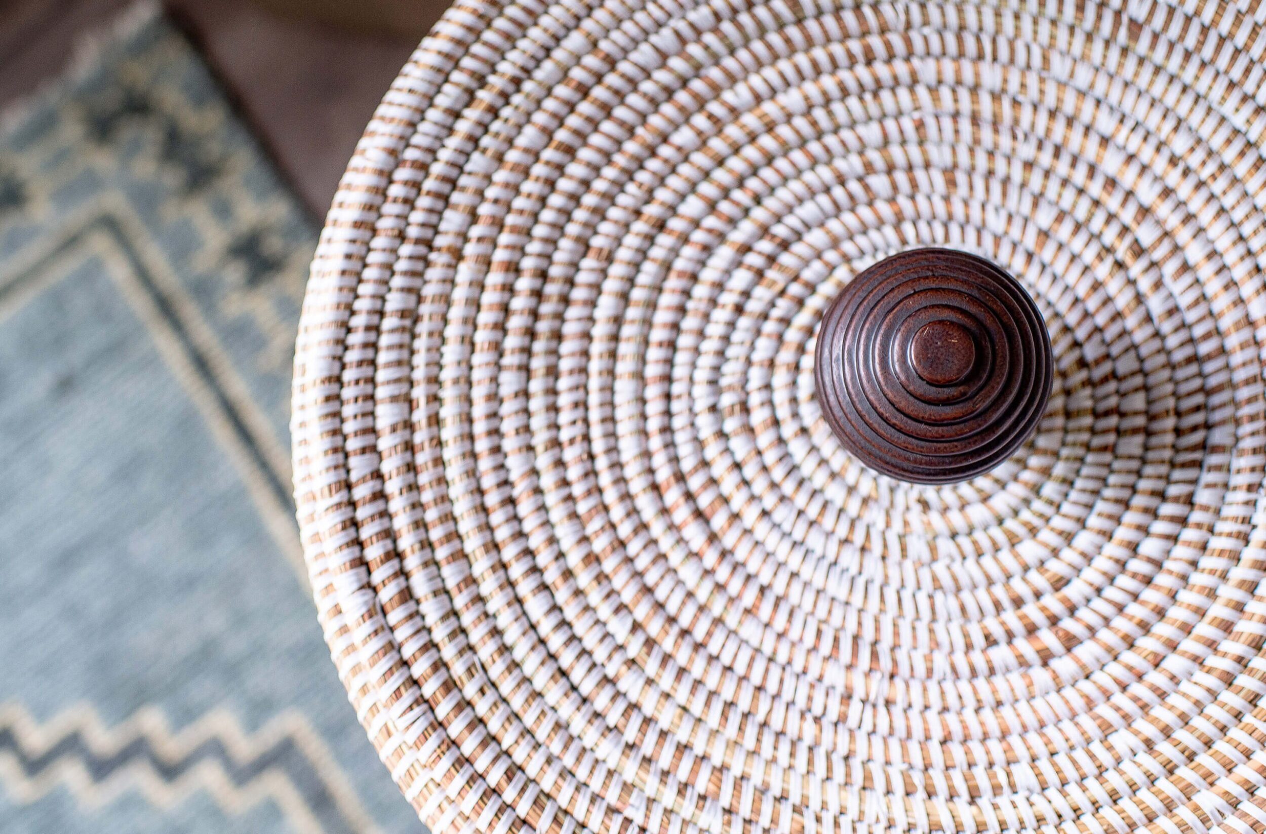 Close-up of woven circular basket lid with wood knob placed on rug, showcasing textured handmade craftsmanship.