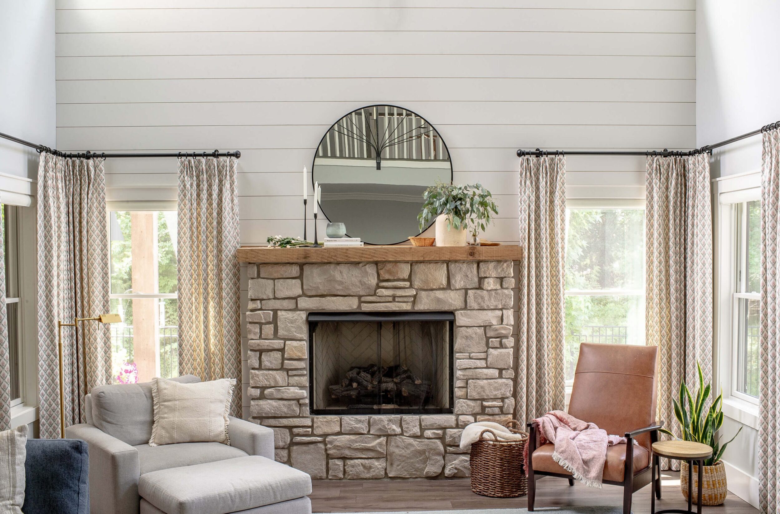 Living room fireplace view with stone surround, wood mantel, round mirror, and cozy seating on both sides.