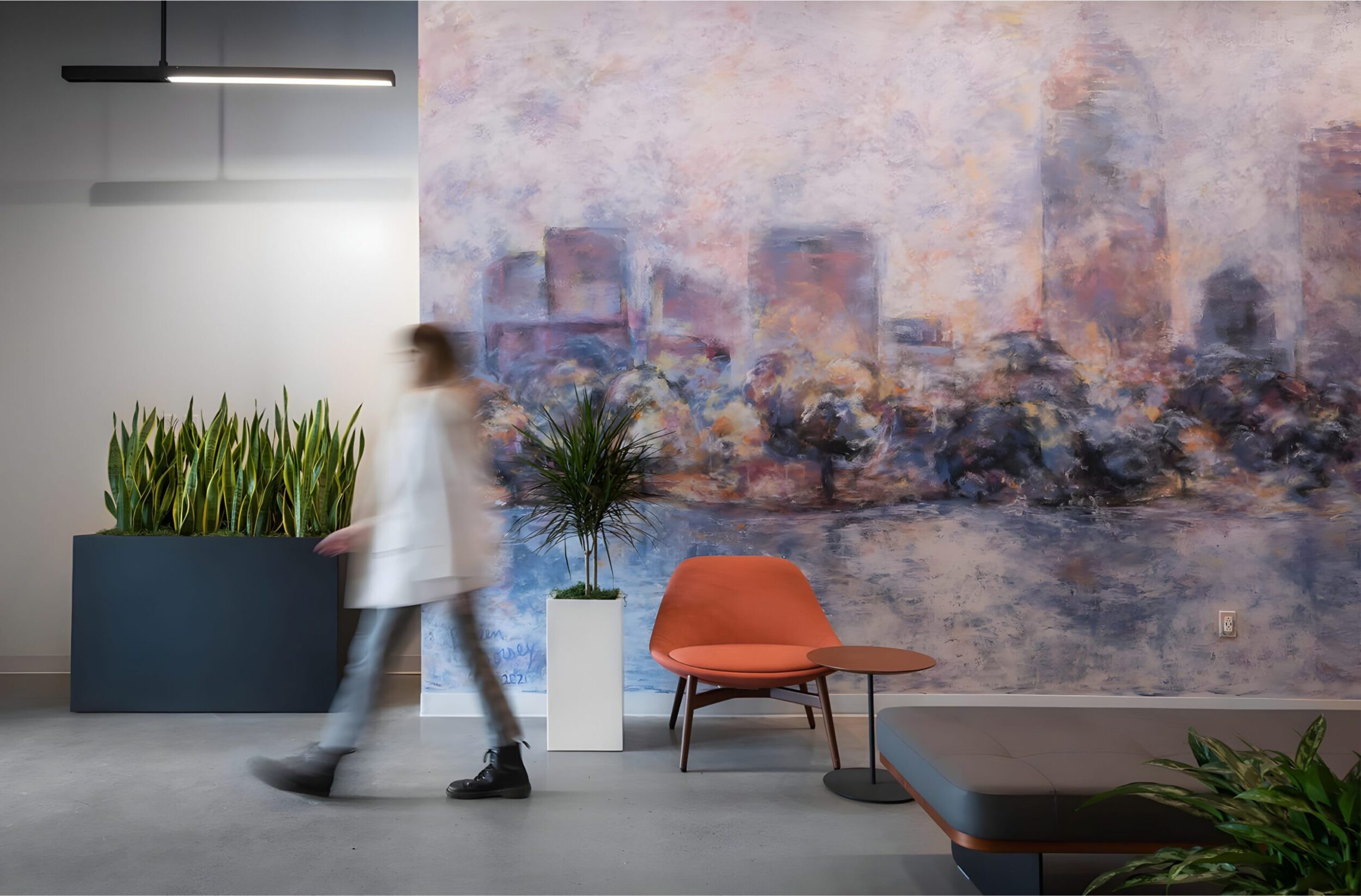 Modern lobby with painted mural, indoor plants, orange chair, and blurred figure walking past.