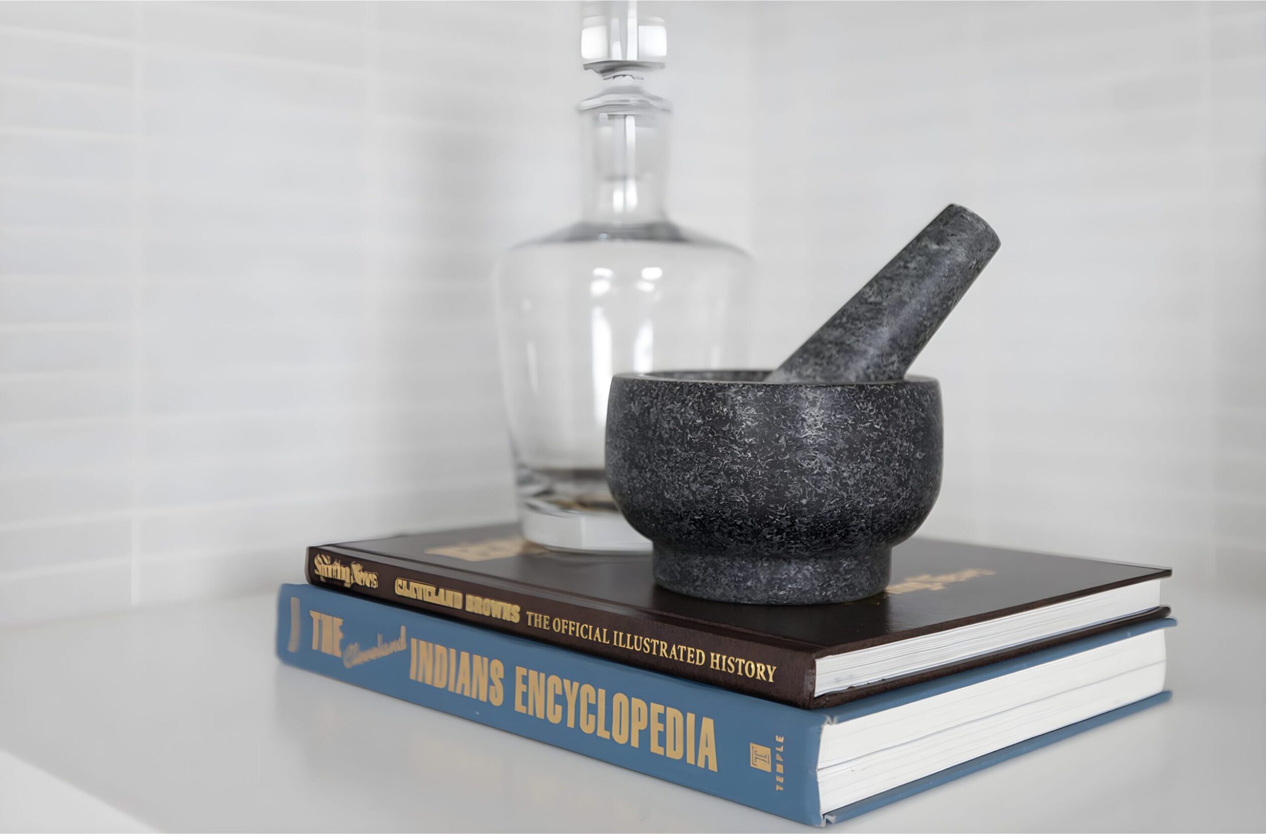 Books stacked with glass decanter and stone mortar and pestle styled on white shelf for modern bar decor.