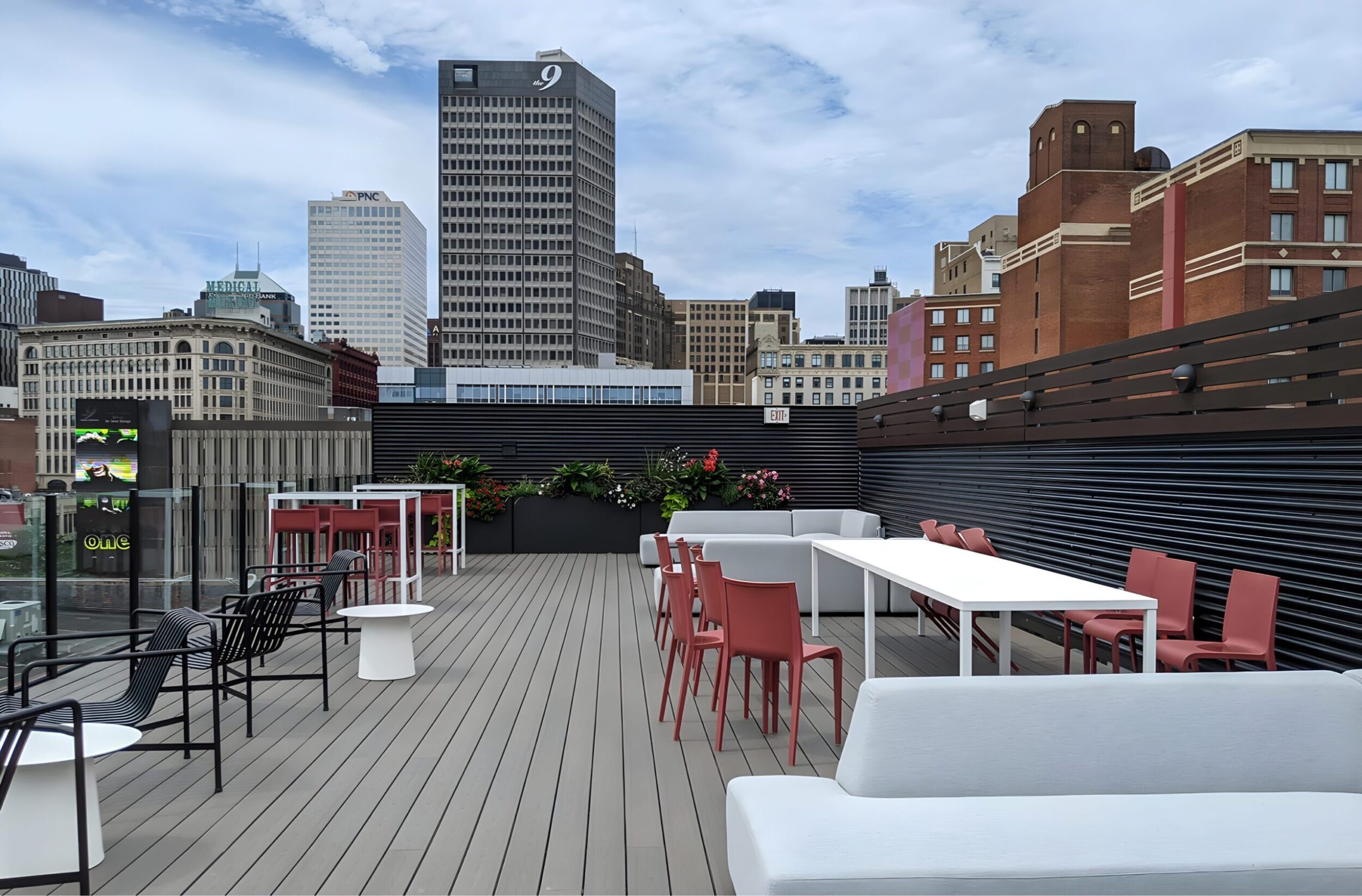 Rooftop deck with modern outdoor seating, city skyline backdrop, and tall buildings under partly cloudy sky.