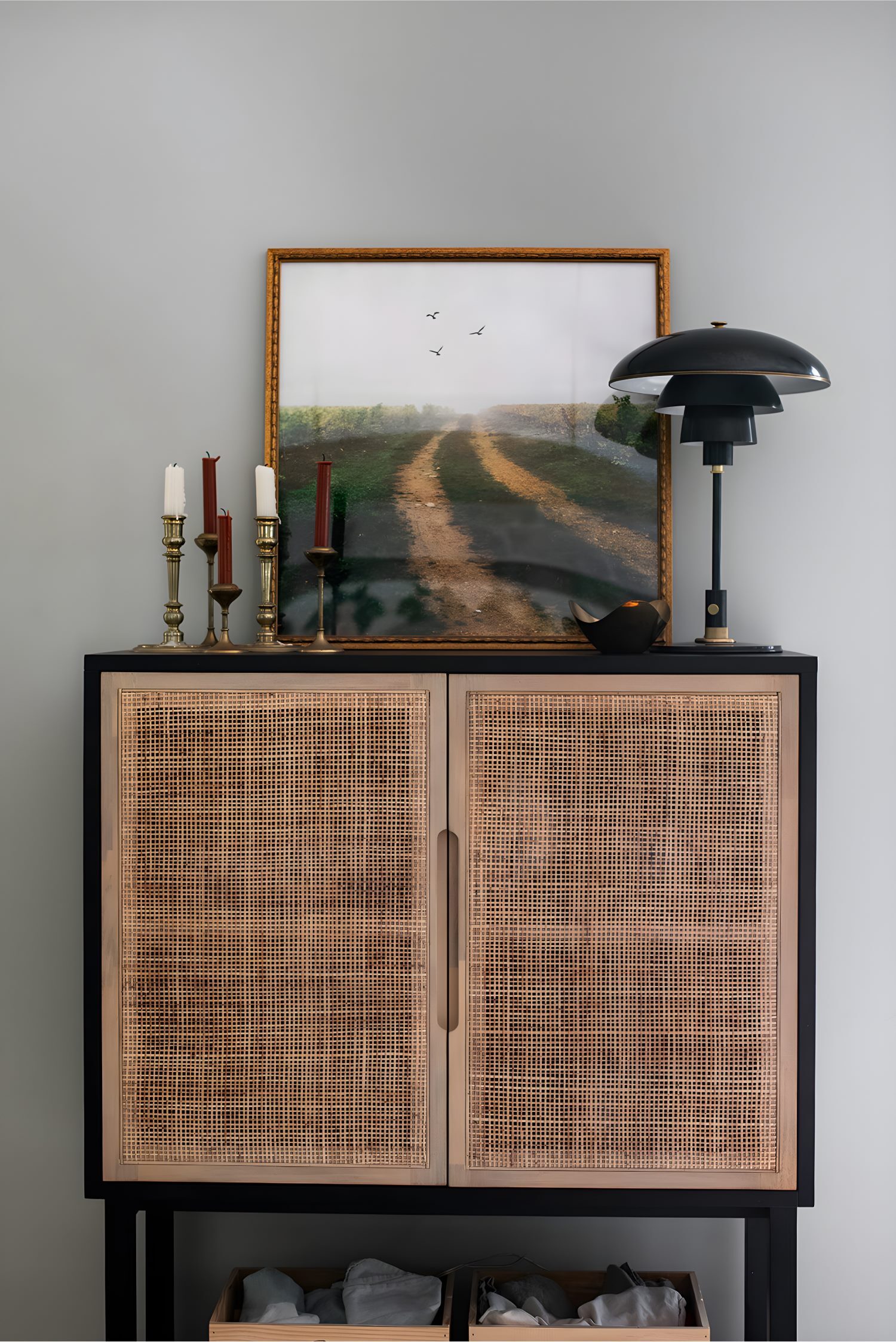 Woven-front cabinet topped with landscape artwork, candlesticks, and modern lamp in a cozy interior vignette.