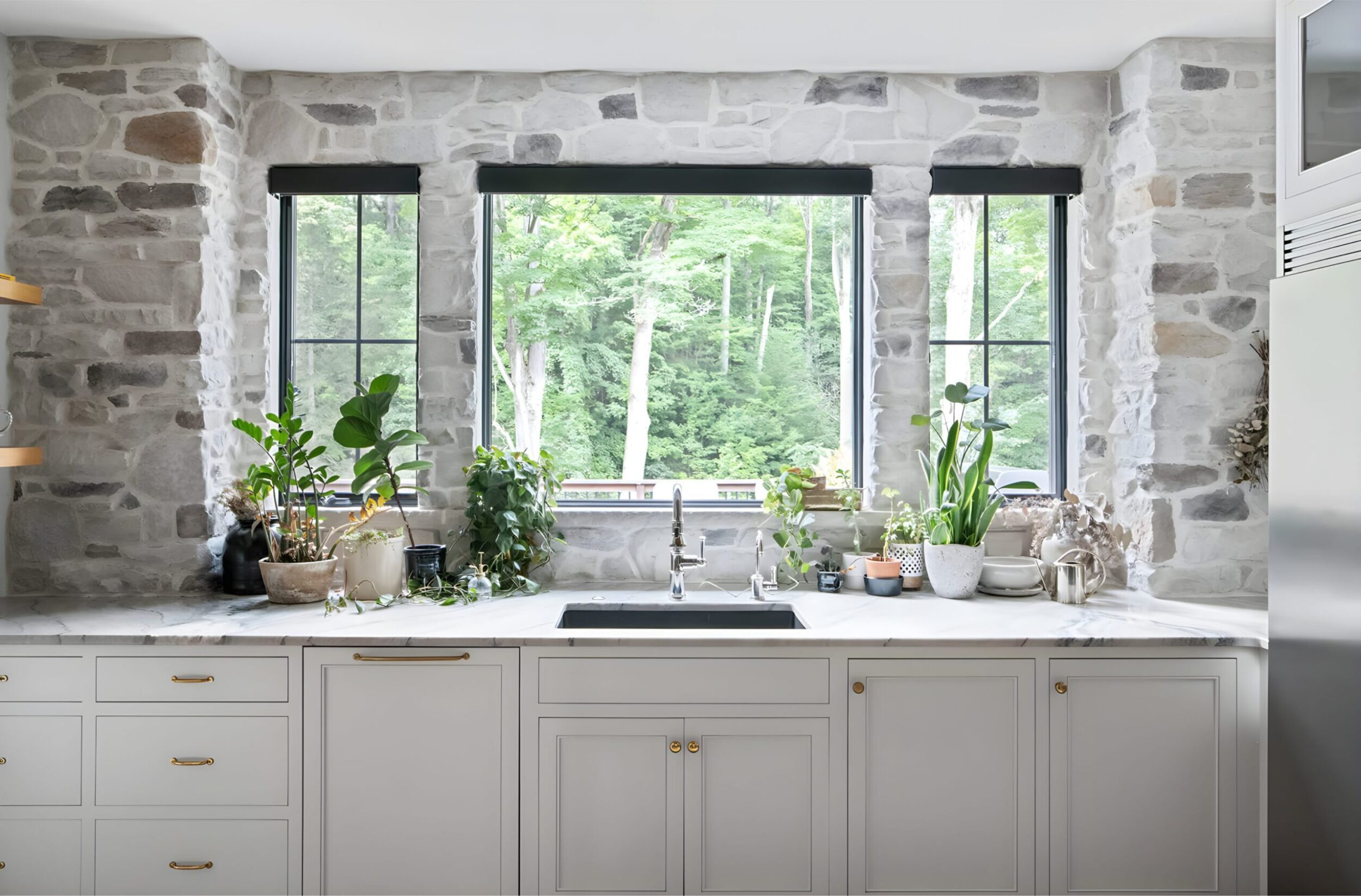 Kitchen sink framed by stone walls and large windows, decorated with potted plants and natural light streaming in.