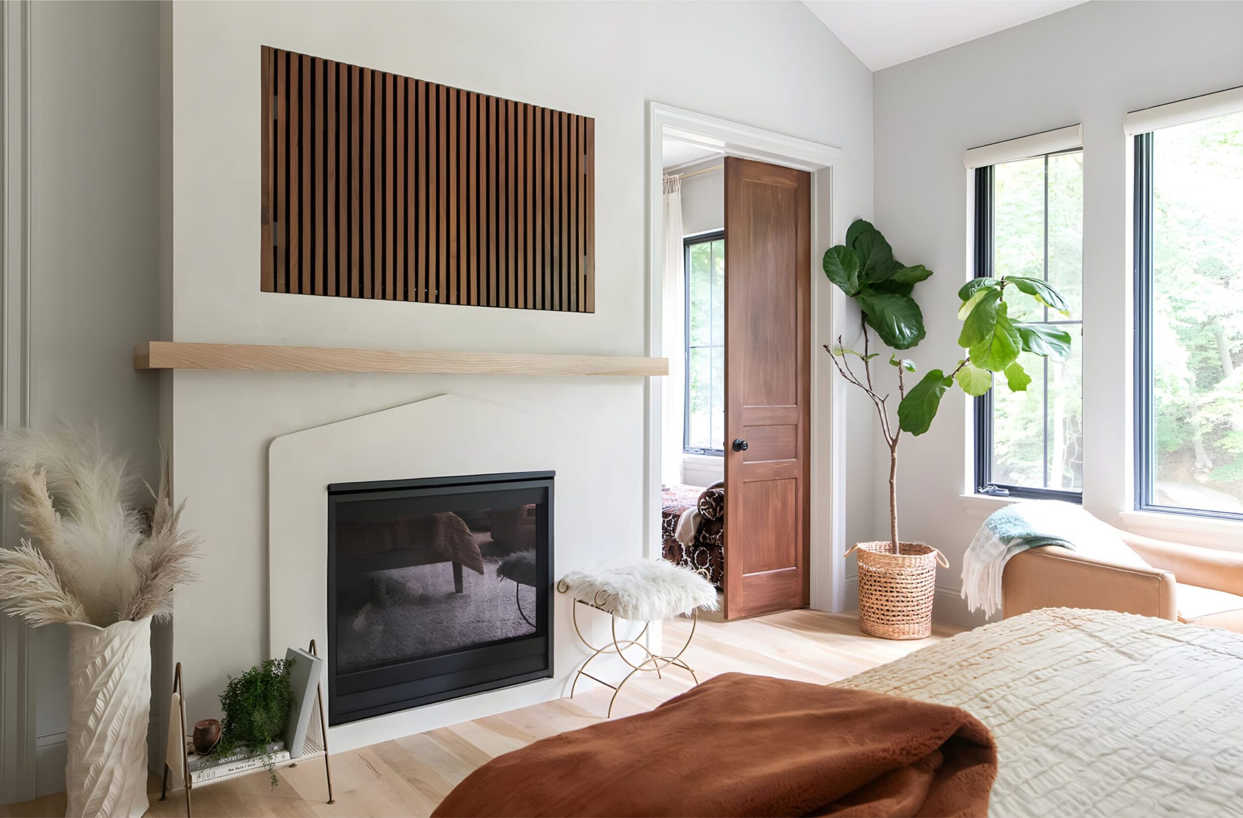 Bedroom with fireplace, wood mantel, woven basket plant, large windows, and cozy layered bedding in warm tones.
