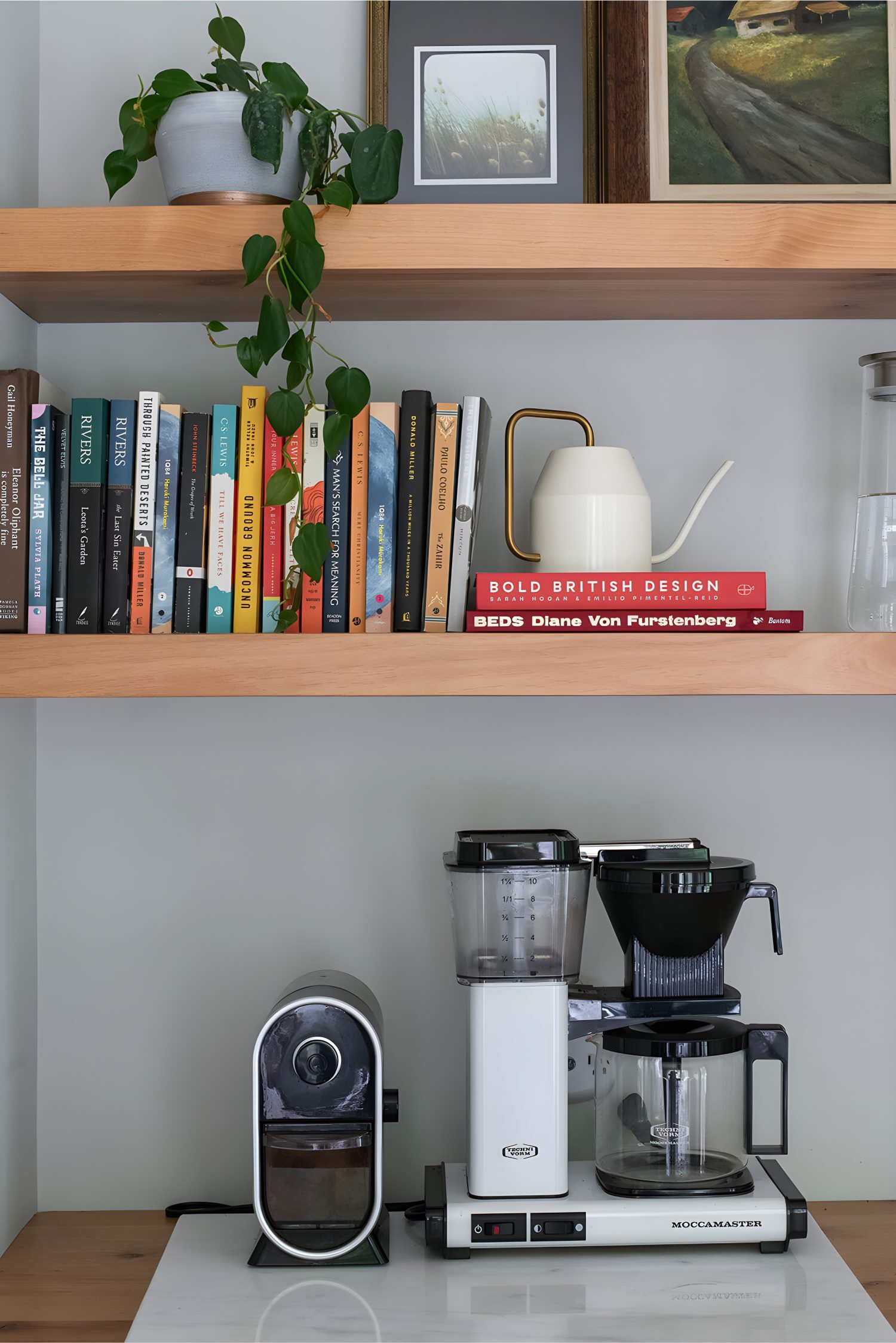 Bookshelf with stacked books, framed art, trailing plant, and coffee machines below on countertop.