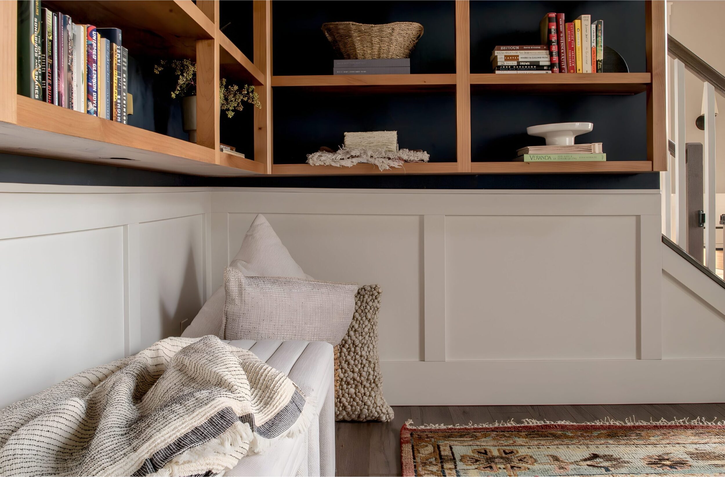 Built-in shelving with books, pillows, and cozy textiles beside staircase, styled with layered cushions and decor.
