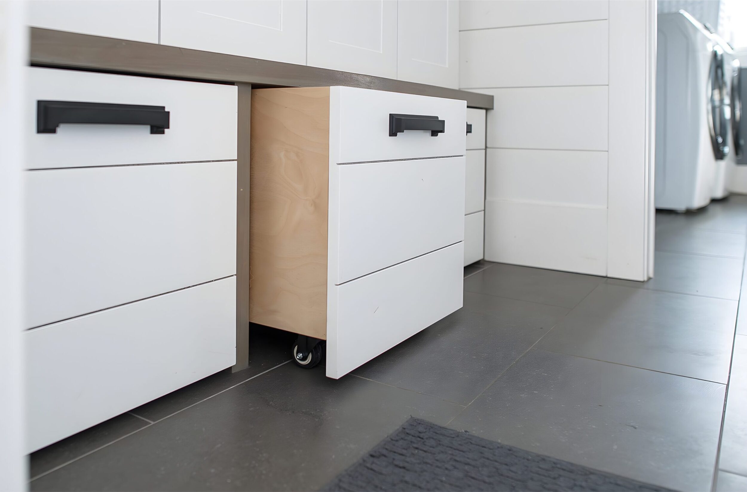 White mudroom cabinetry with black handles, showing open drawer on casters for hidden storage in tiled hallway.