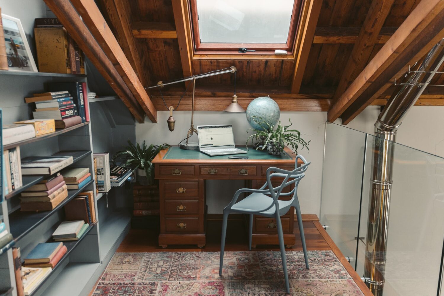 Attic office space with vintage wooden desk, laptop, bookshelves, globe, and skylight window.