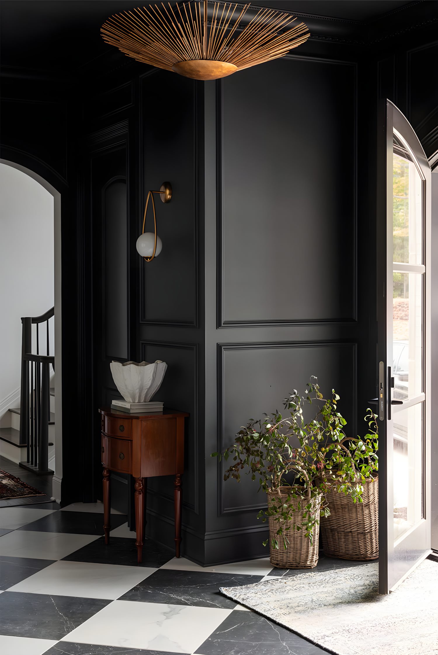 Entryway with black paneled walls, gold sunburst ceiling light, wicker baskets of greenery, and black and white checkered floor