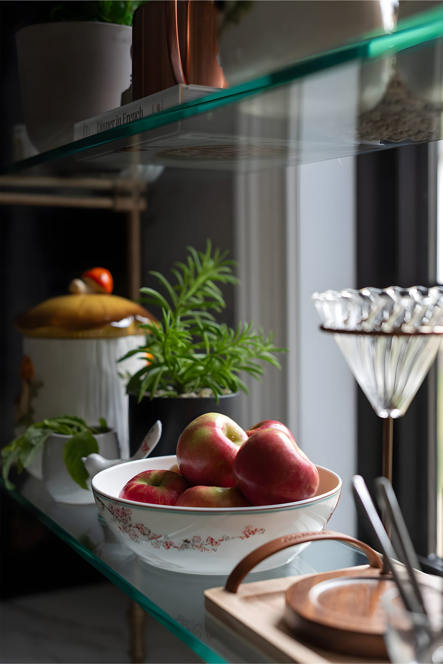 Close-up of glass shelf with white floral bowl of apples, copper pitcher, plants, and clear martini glass in natural light