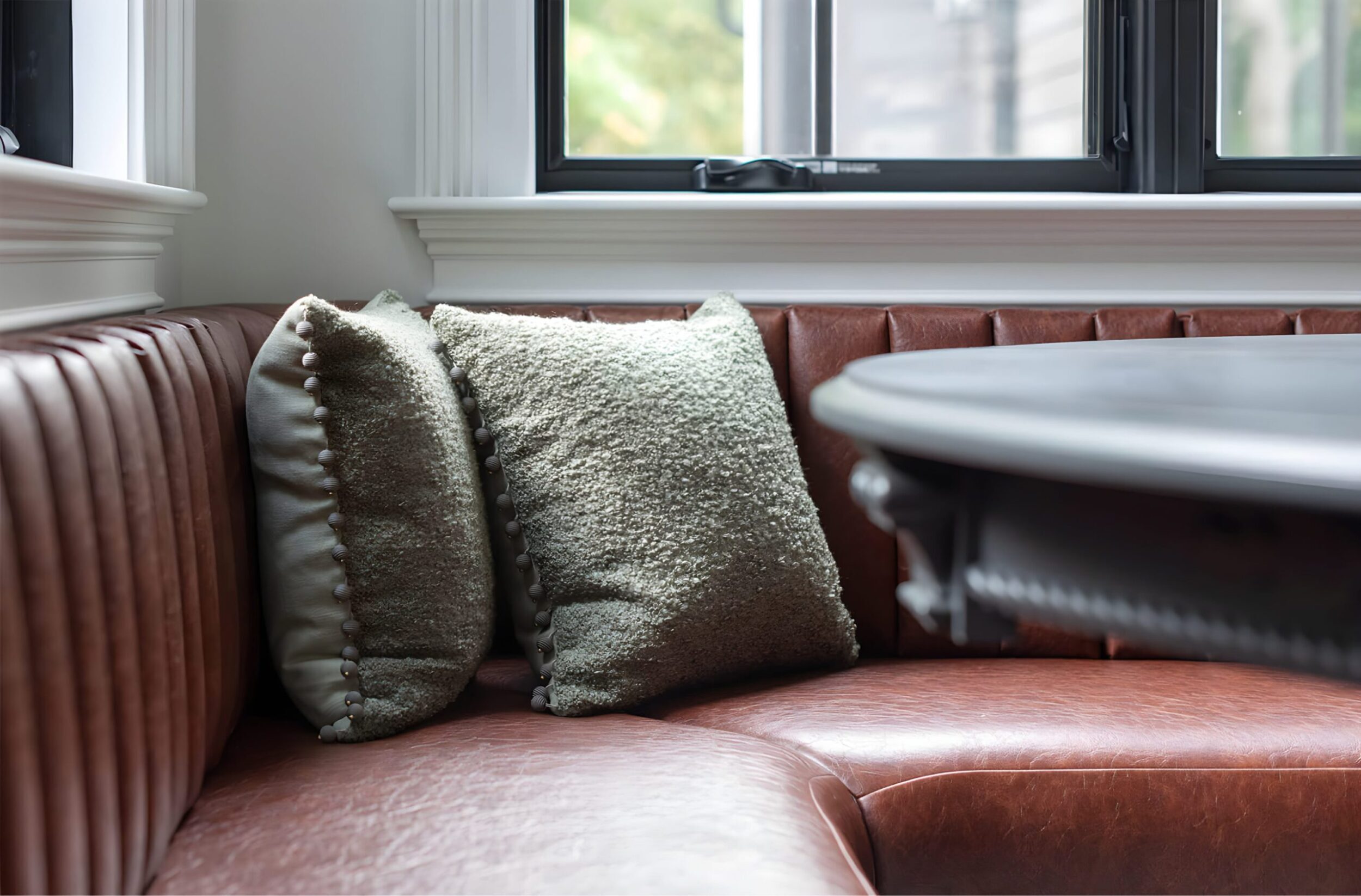 Brown leather banquette seating with vertical tufting, styled with textured green accent pillows by a black round table