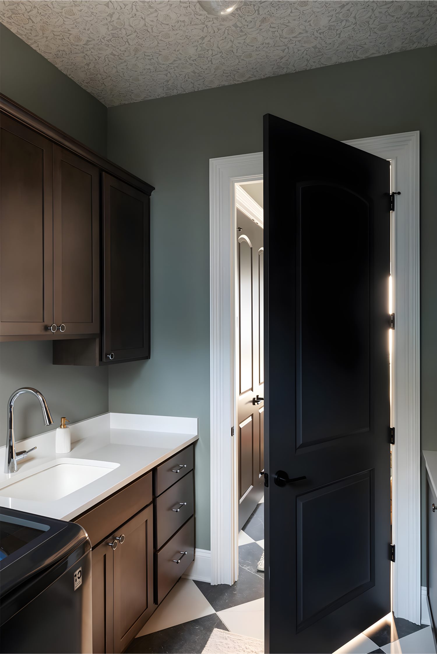 Laundry room with dark cabinets, white countertop, stainless faucet, patterned ceiling, and black door ajar
