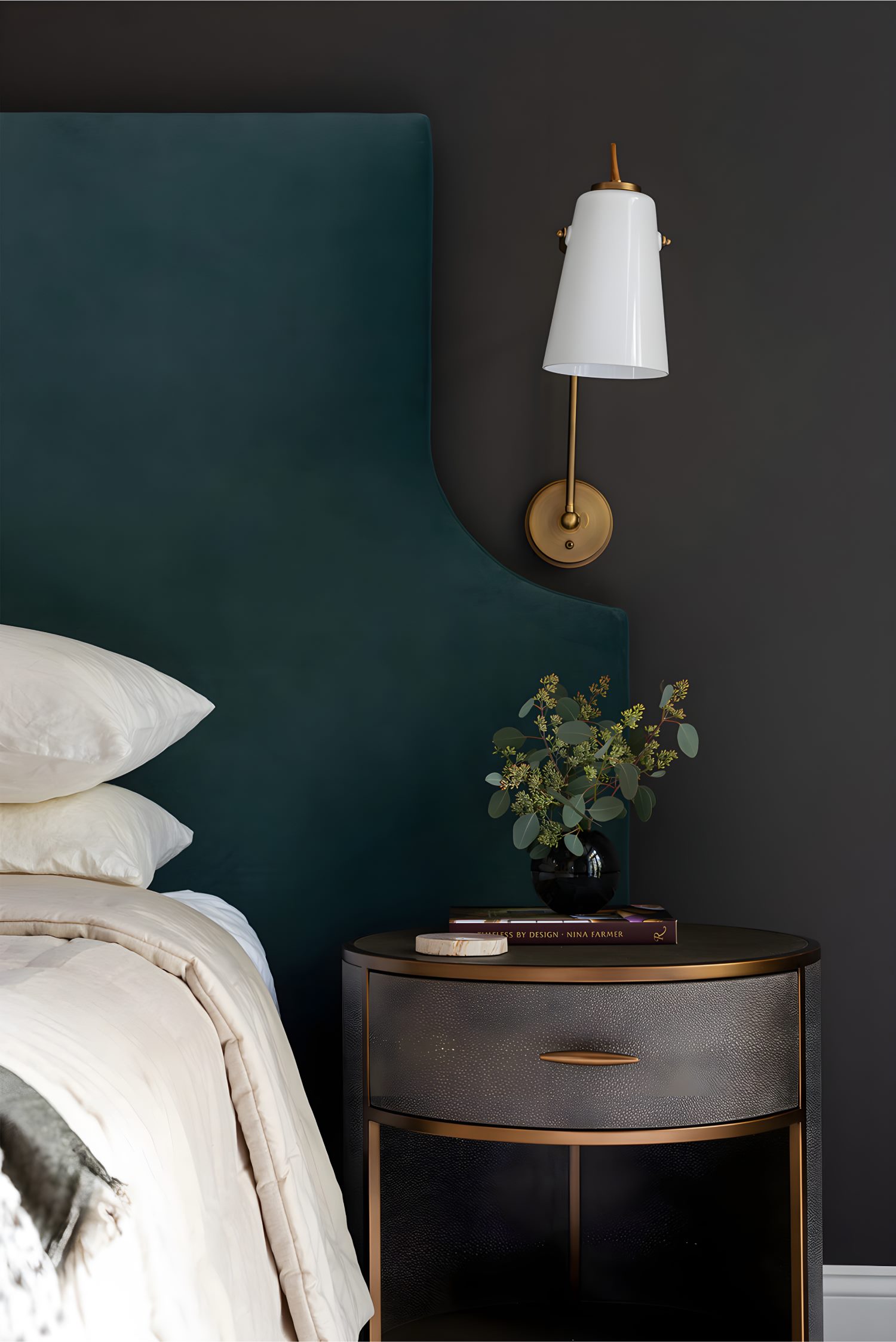 Bedside table with round drawer, brass lamp, black vase with greenery, books, and teal headboard in background