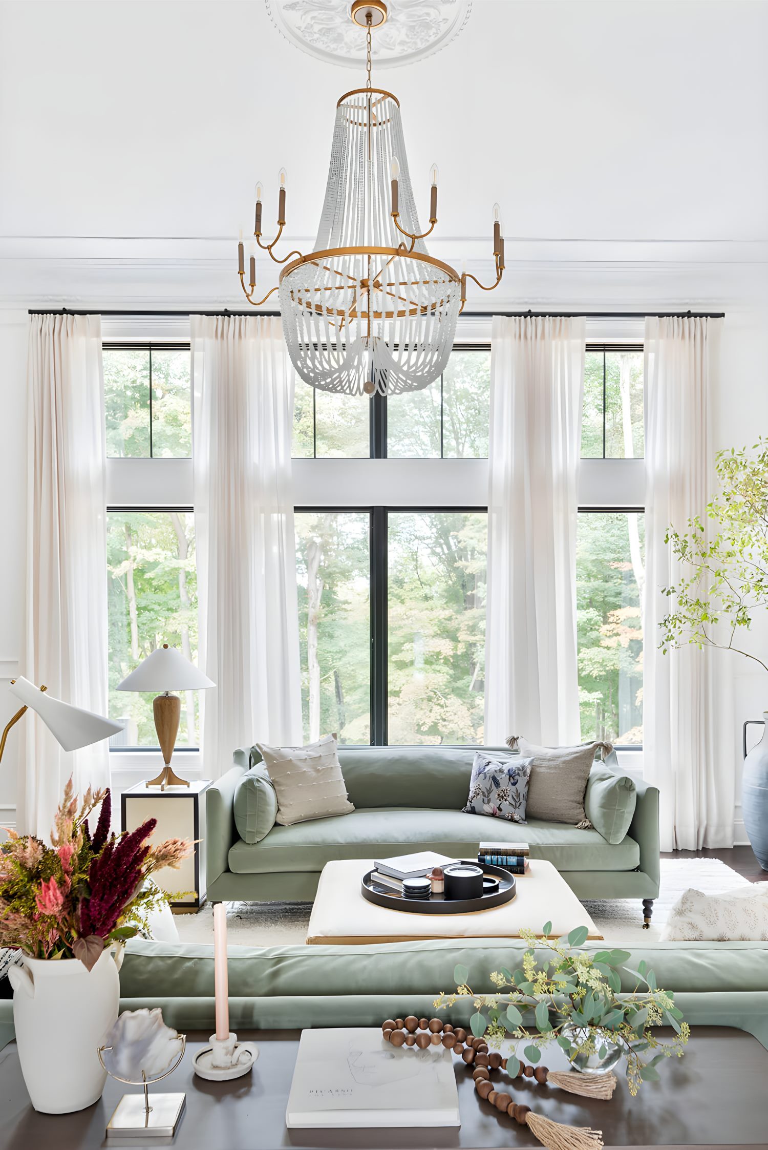 Living room with tall windows, soft green sofas, chandelier, sheer curtains, and modern ottoman coffee table
