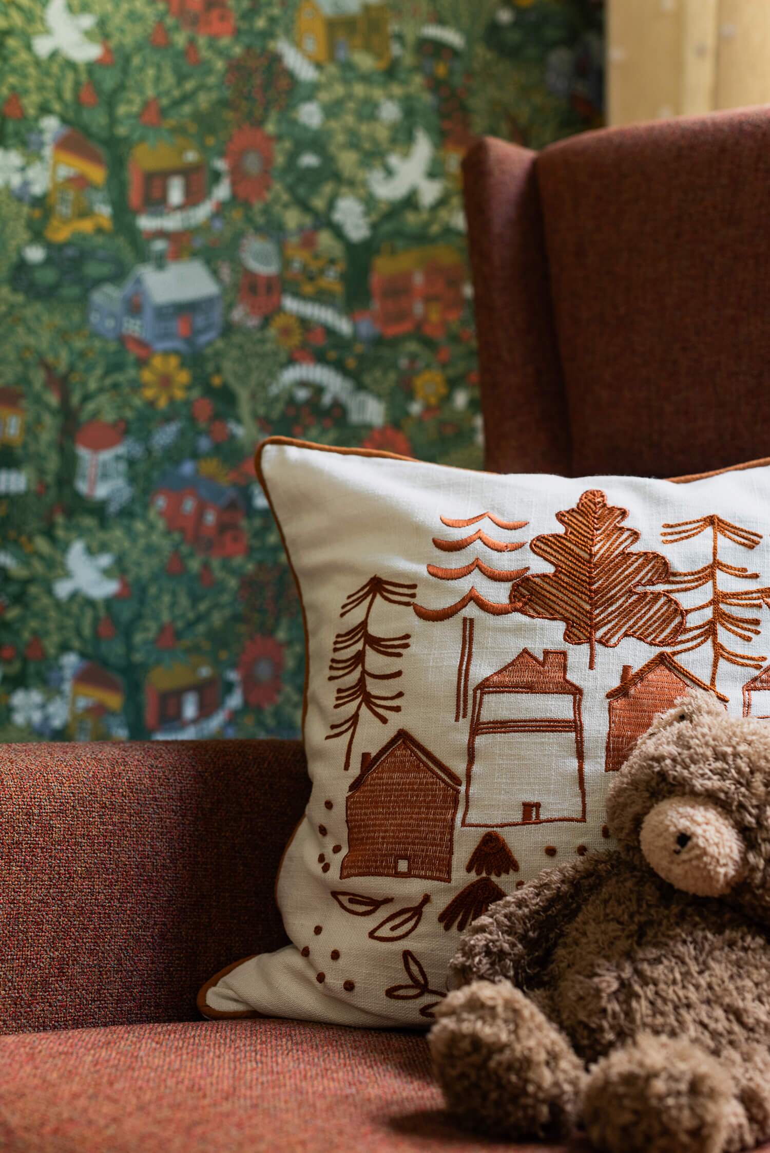 Rust-toned armchair holds embroidered nature-scene cushion and plush teddy; vibrant botanical wallpaper with houses behind.Rust-toned armchair holds embroidered nature-scene cushion and plush teddy; vibrant botanical wallpaper with houses behind.