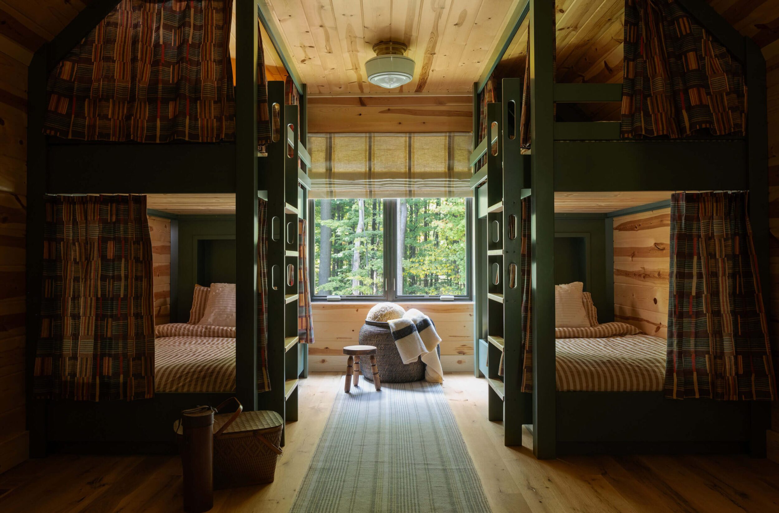 Rustic bunk room with twin bunks framed in olive-green, patterned curtains, central light, cozy rug, stool, and forest window view.