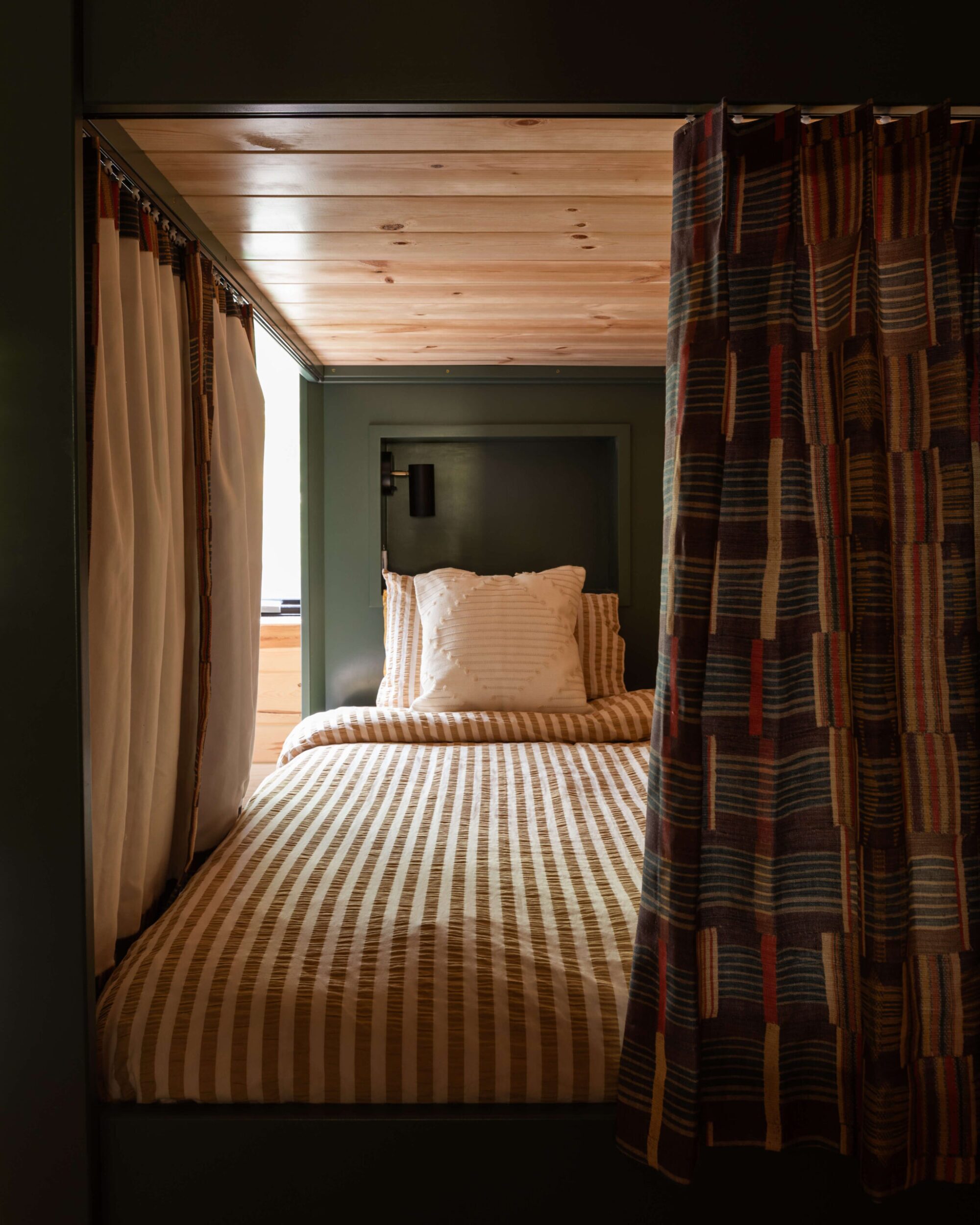 Cozy bunk with striped beige bedding, wood-paneled ceiling, forest-green built-in frame; layered curtains create a private nook.