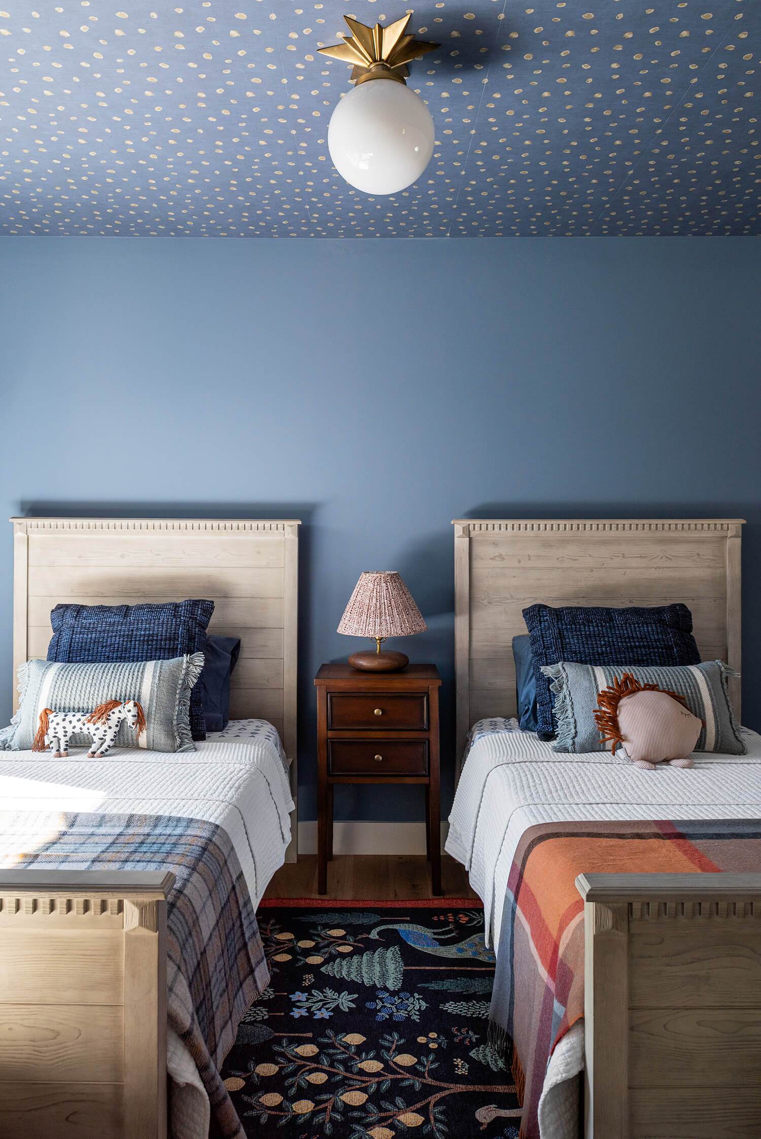Twin beds with blue accent wall, matching headboards, patterned rugs, and playful pillows in kids’ room.