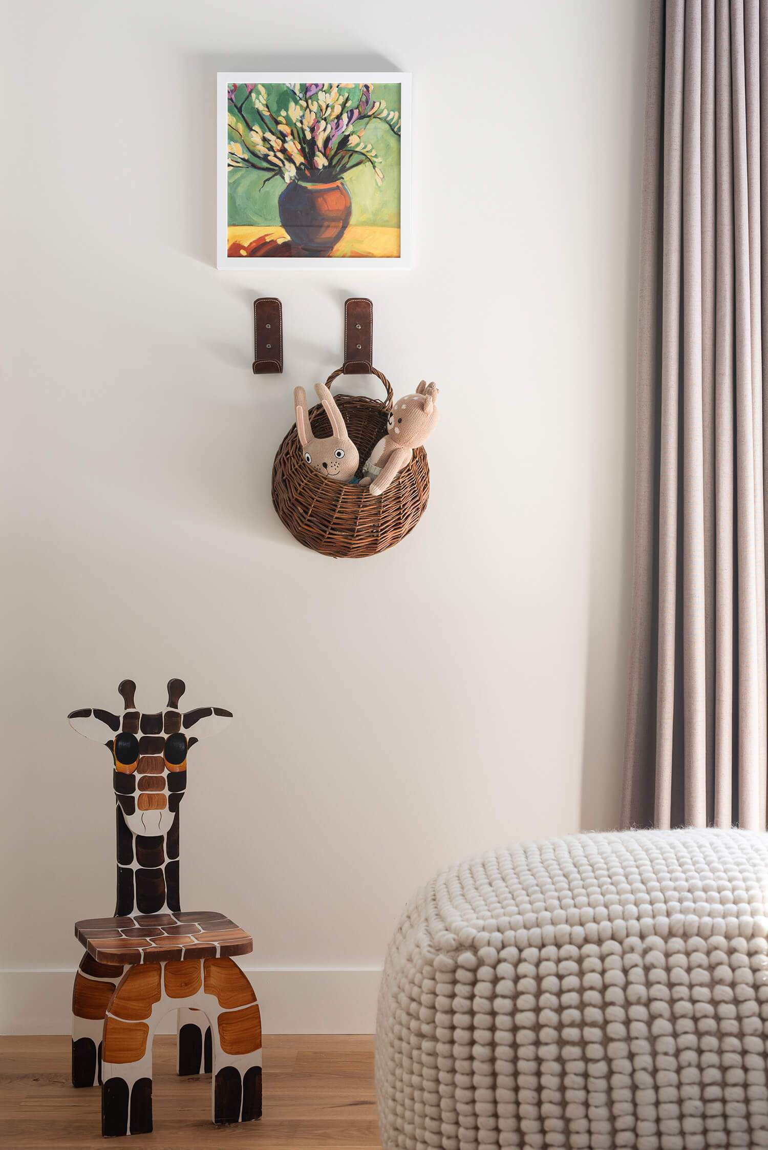 Wall hooks with basket of plush toys, giraffe chair, and framed floral artwork above in kids’ bedroom.