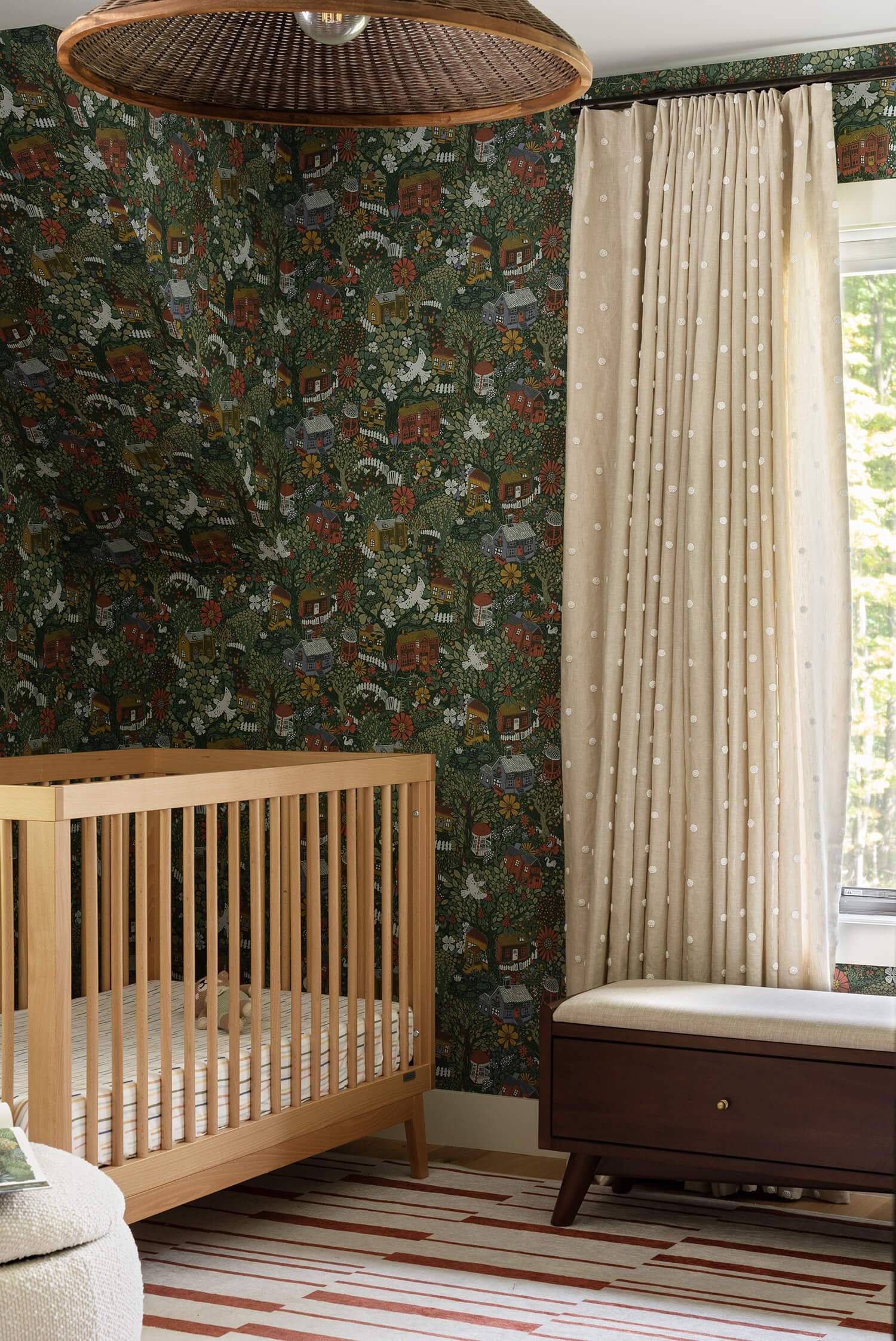 Light wood crib on patterned rug in nursery with detailed green botanical wallpaper, beige dotted drapes, wicker pendant lamp.