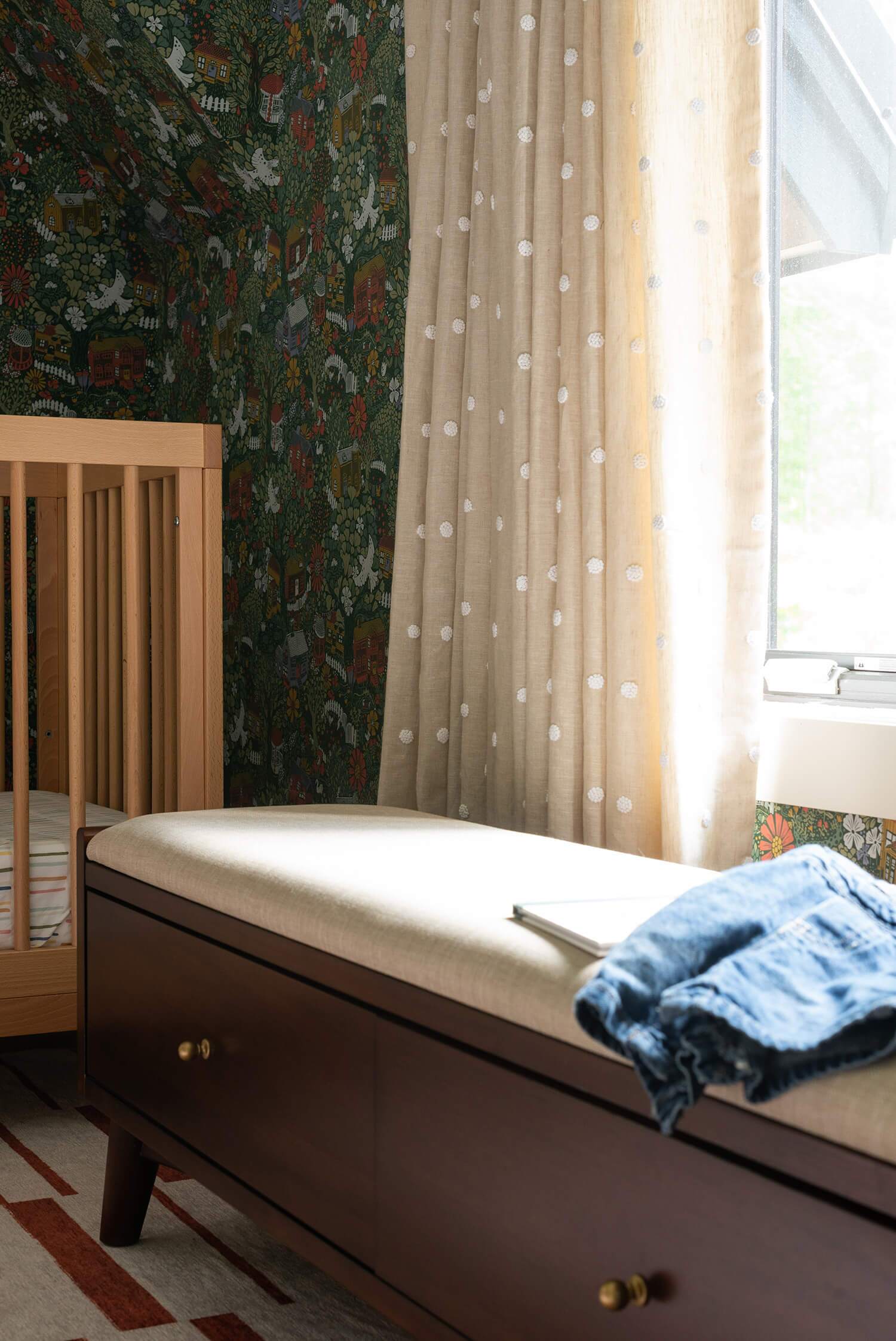 Wooden crib beside cushioned bench with drawer, blue blanket; polka-dot beige drapes over botanical wallpaper by a sunlit window.
