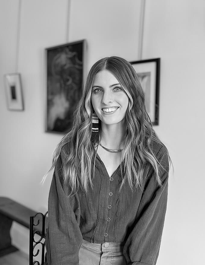Black and white portrait of smiling woman with long hair, wearing buttoned shirt, seated with framed art in background