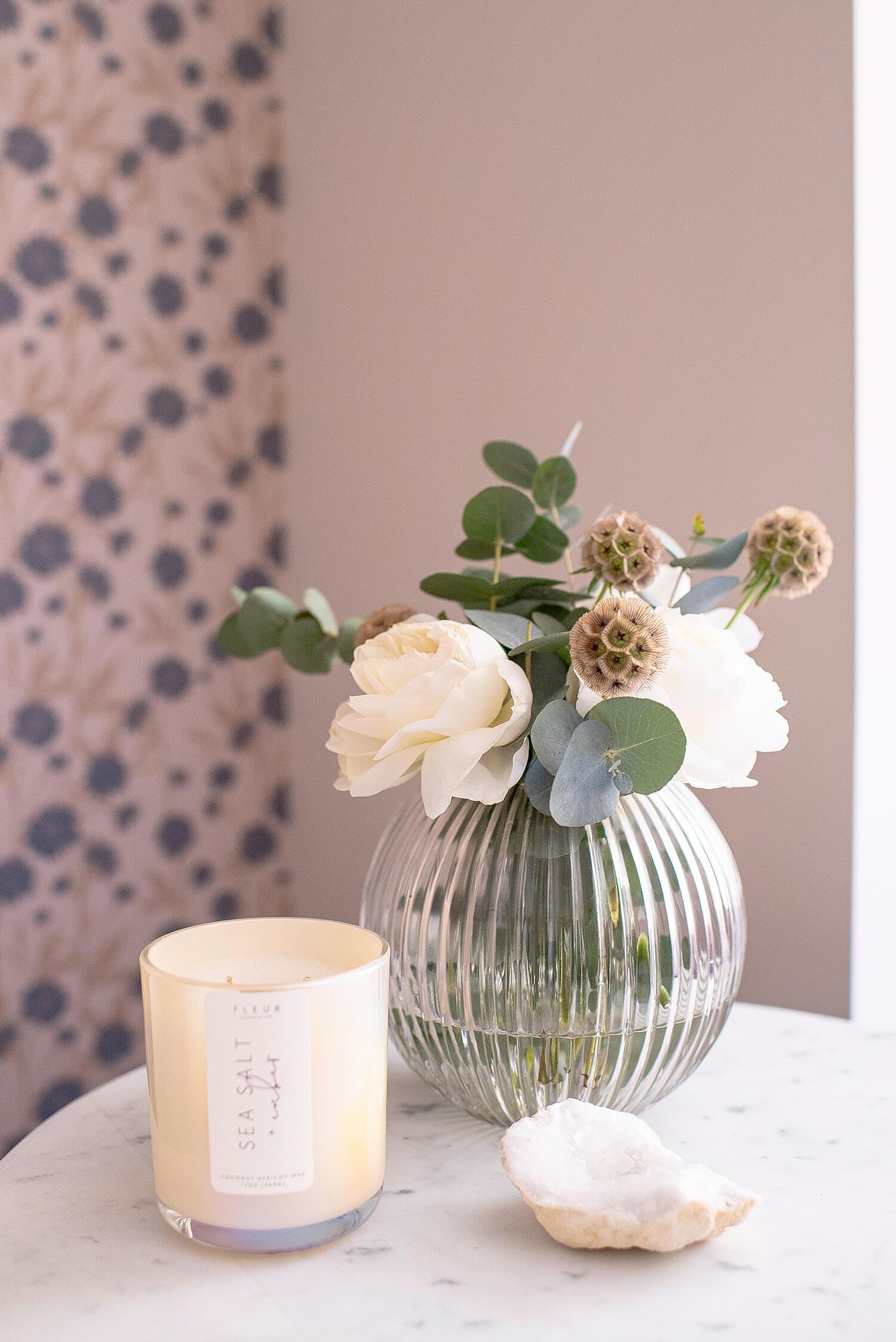 Glass vase with white roses and eucalyptus beside candle and crystal decor on marble table against floral wallpaper accent.