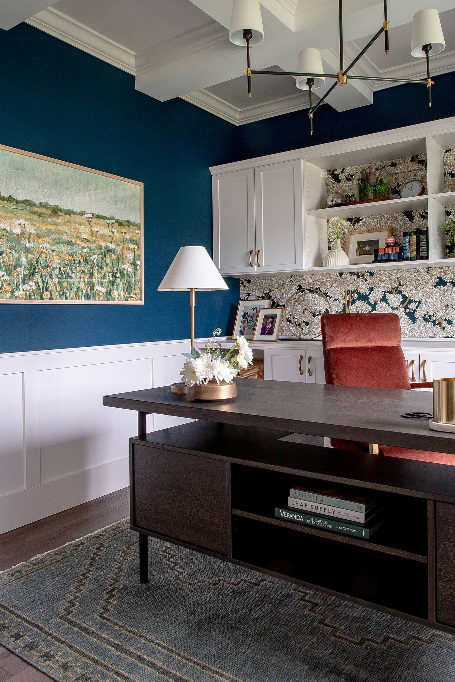 Home office with navy walls, floral wallpaper, white cabinetry, and coral chair styled with books and framed art.