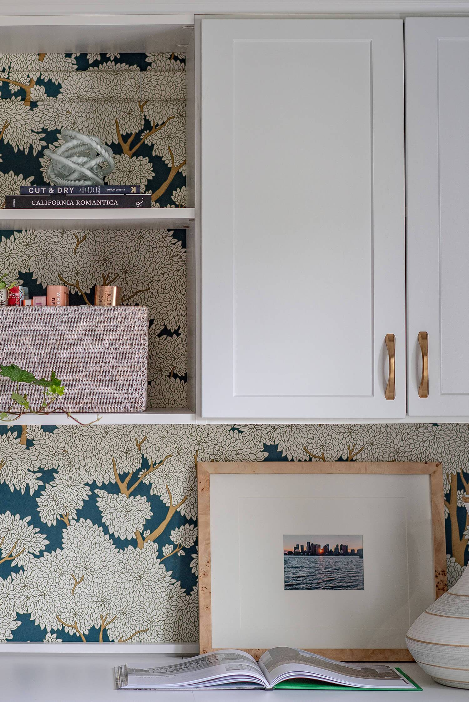 White built-in cabinetry with floral wallpaper, framed art, decor items, and open book styled for interior design.